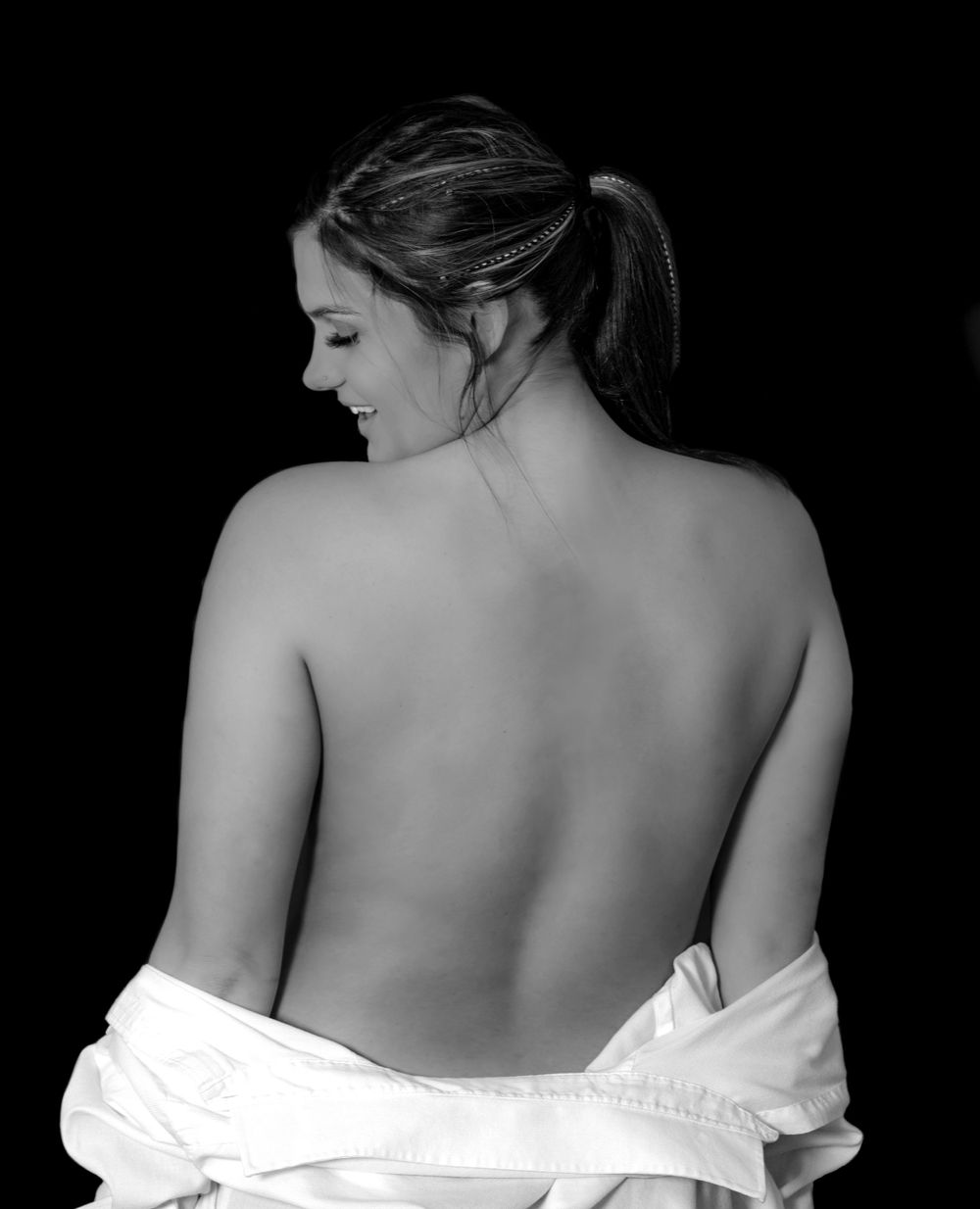 This glamour shot in black and white shows a young woman with a bare back as her shirt man's shirt is around her waist.