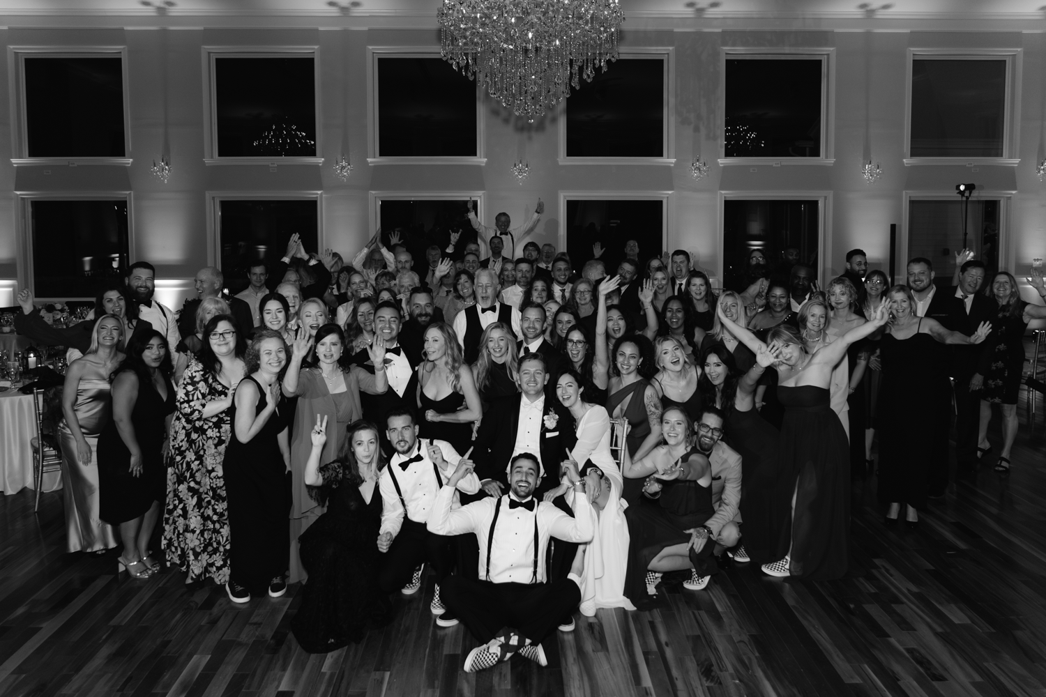 Large group photo of wedding guests gathered on a dance floor under crystal chandeliers.
