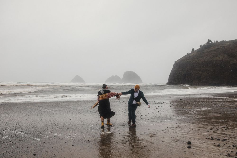 Two people holding hands while walking along a misty beach with sea stacks in the background.