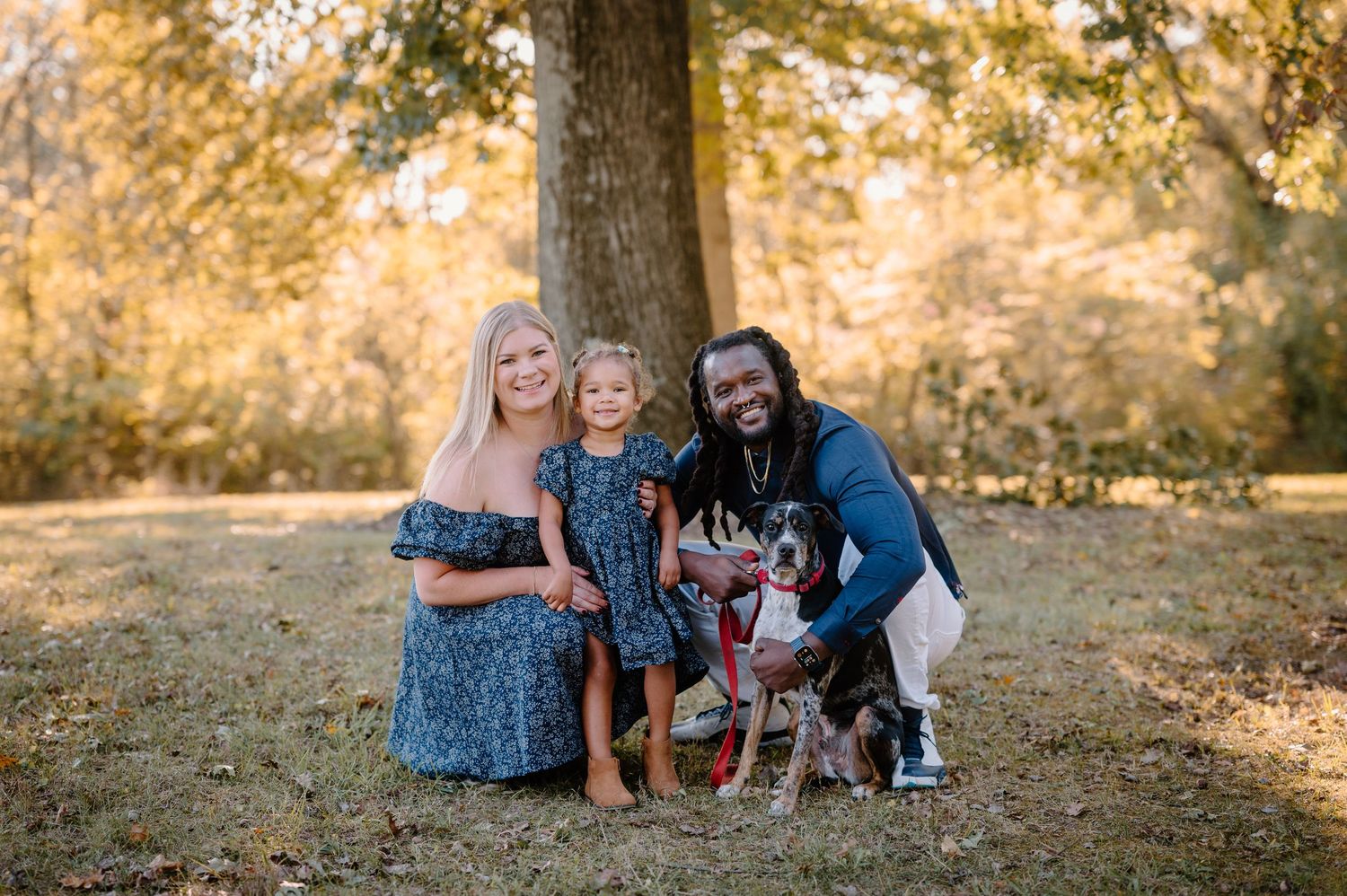 A smiling family poses together outdoors with their dog on an autumn day under a large tree.