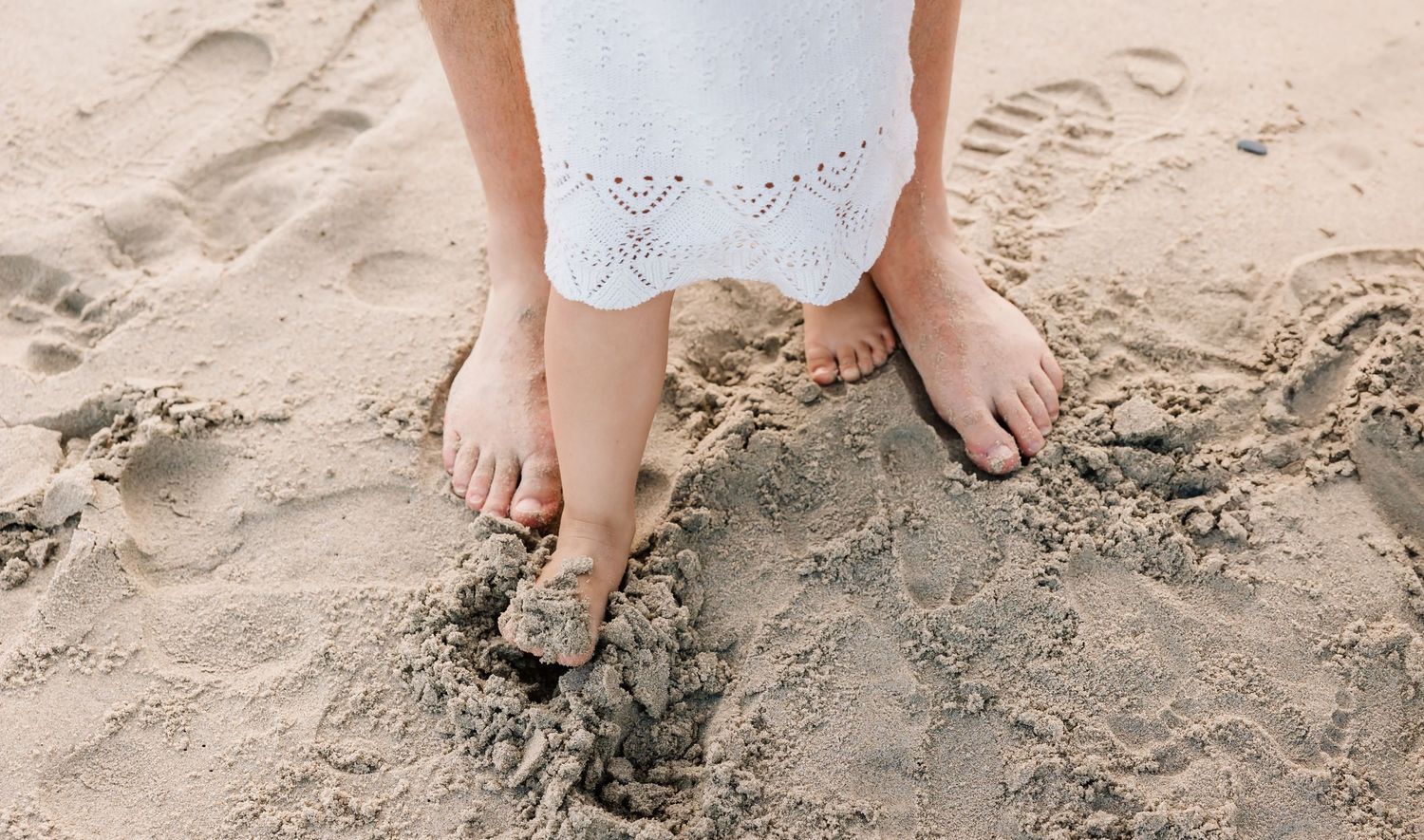 Bare feet in a white lace dress stand on sandy beach with footprints in the sand.