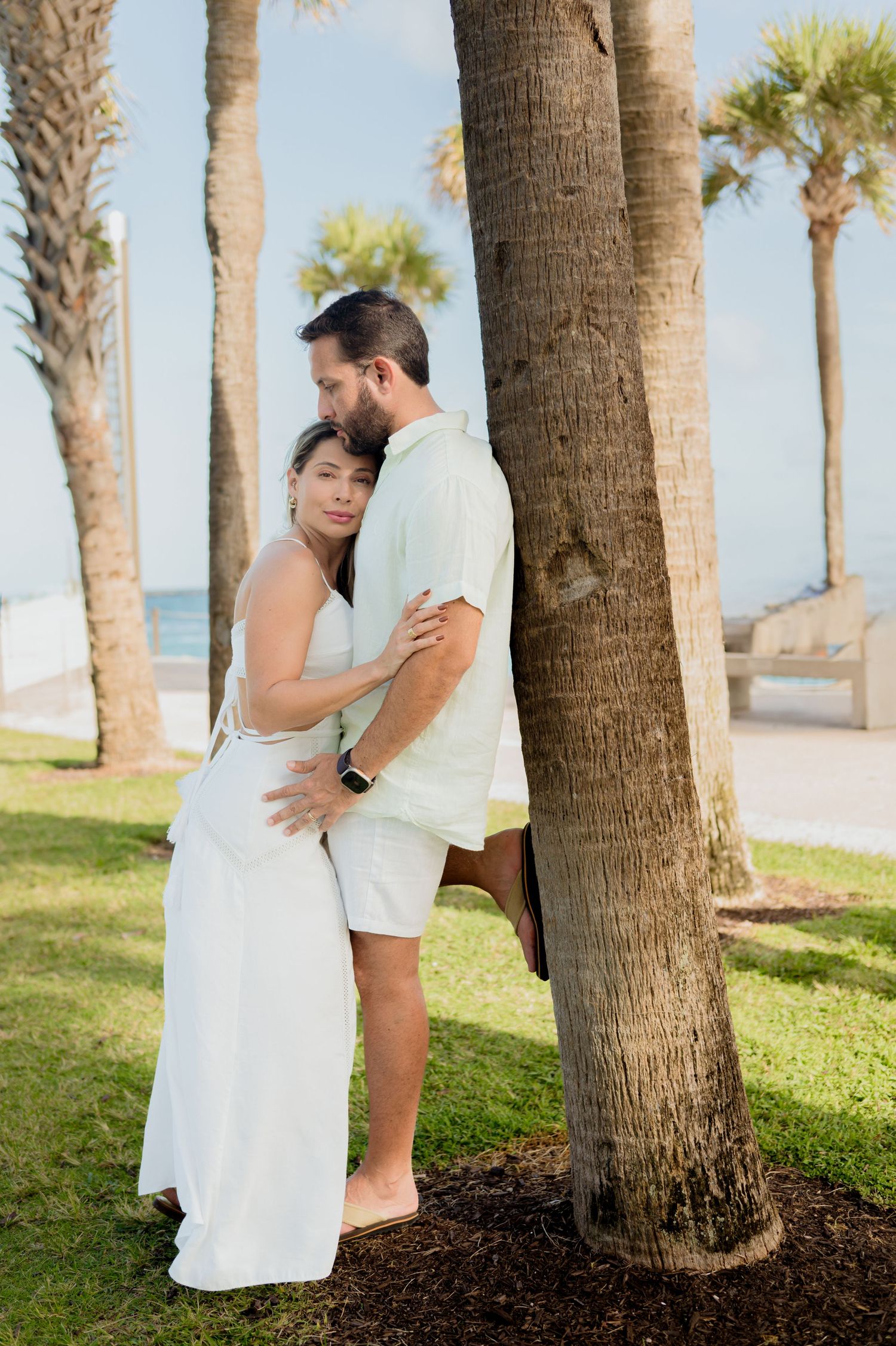 A romantic couple in white outfits embraces by palm trees on a tropical beach during a sunset photoshoot.