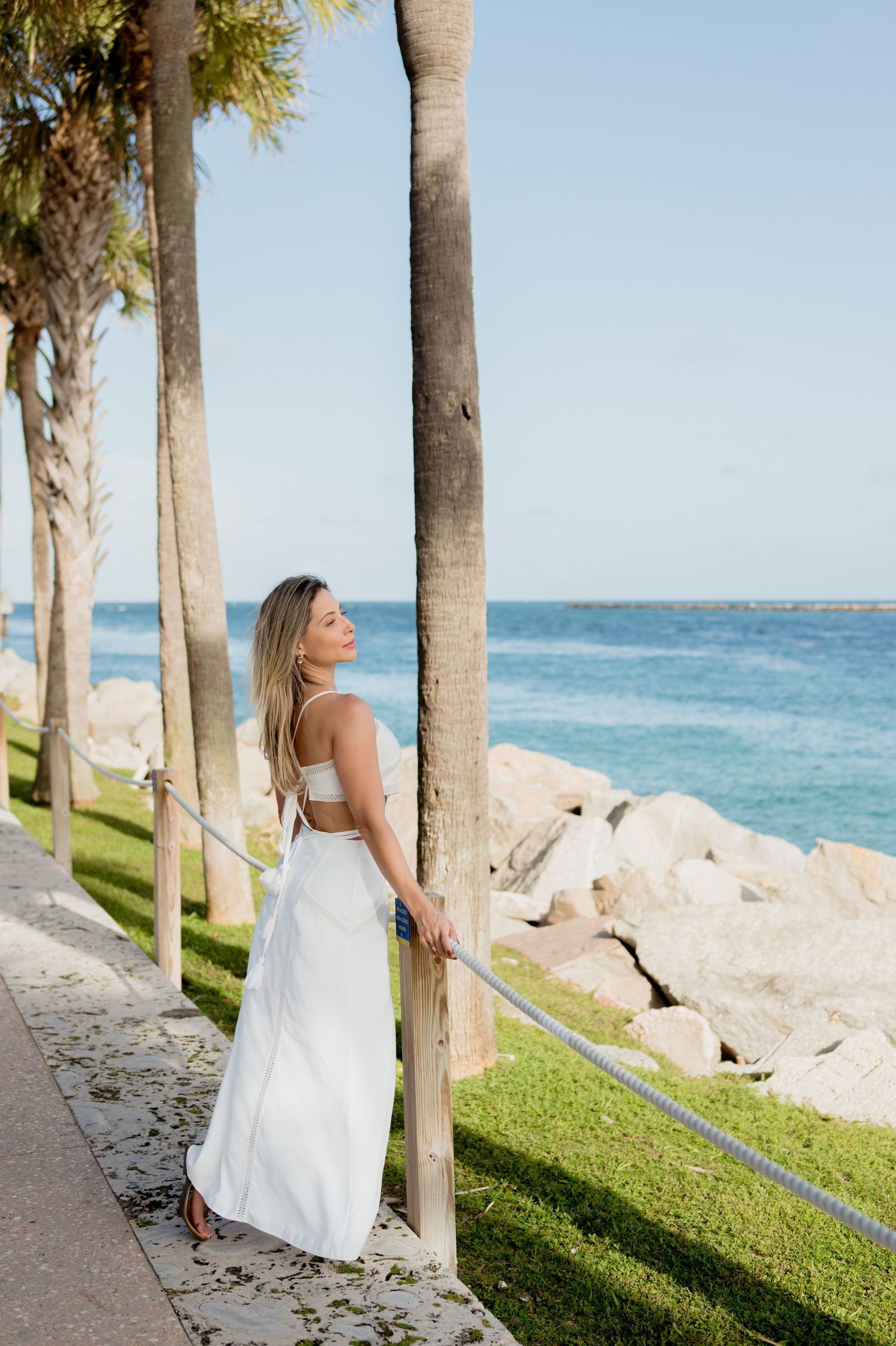 A woman in a white dress poses along a seaside path with palm trees and a rocky shoreline in the afternoon sun.