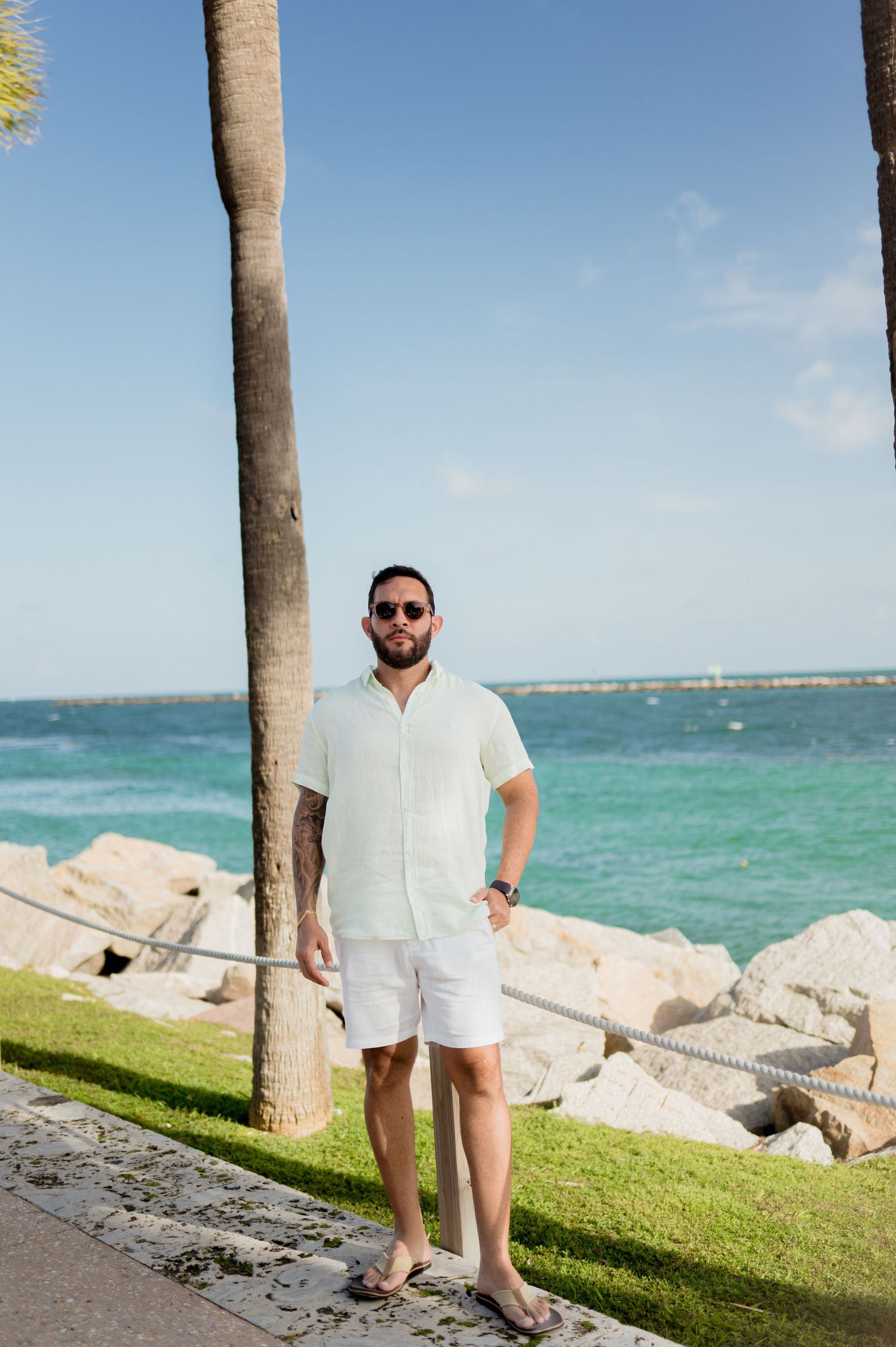 Person in casual summer attire stands near palm tree on rocky beach with turquoise ocean waters in background.