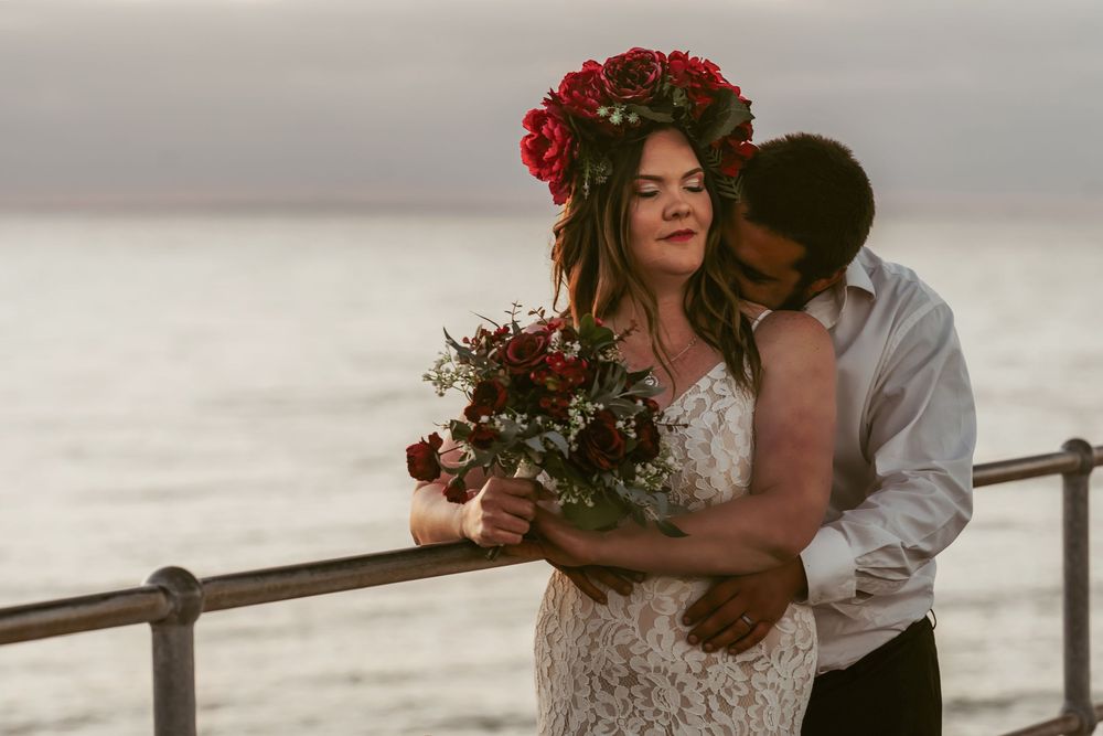 Two people share intimate moments in formal wedding attire on a beachside deck at sunset, with red floral bouquet decorations.