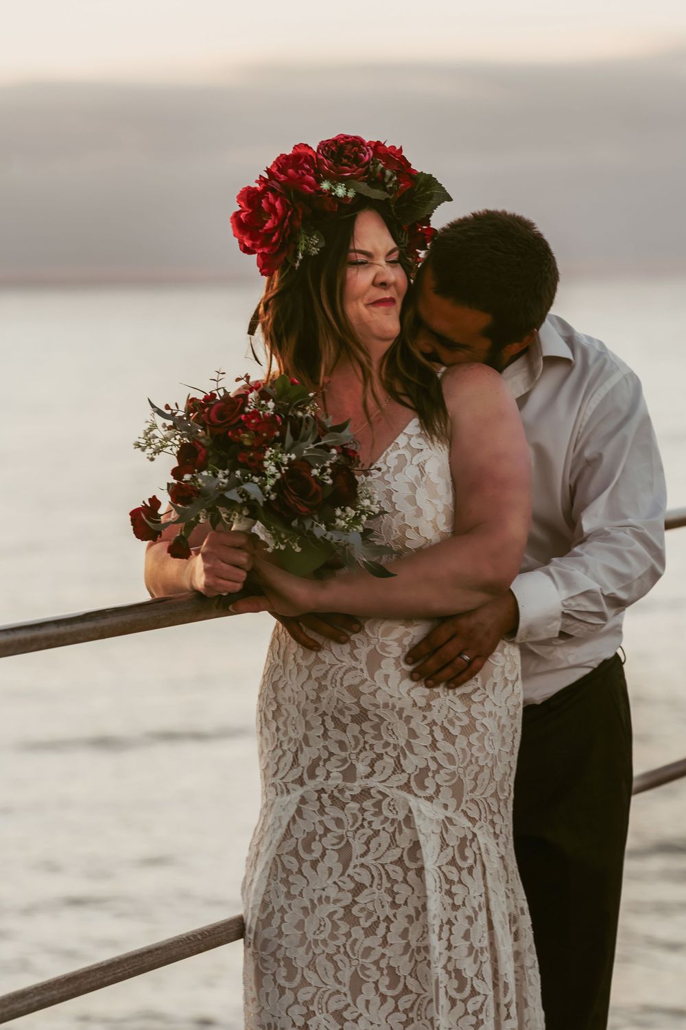 A romantic wedding couple embraces by the water at sunset, with the bride wearing a lace gown and red flower crown.