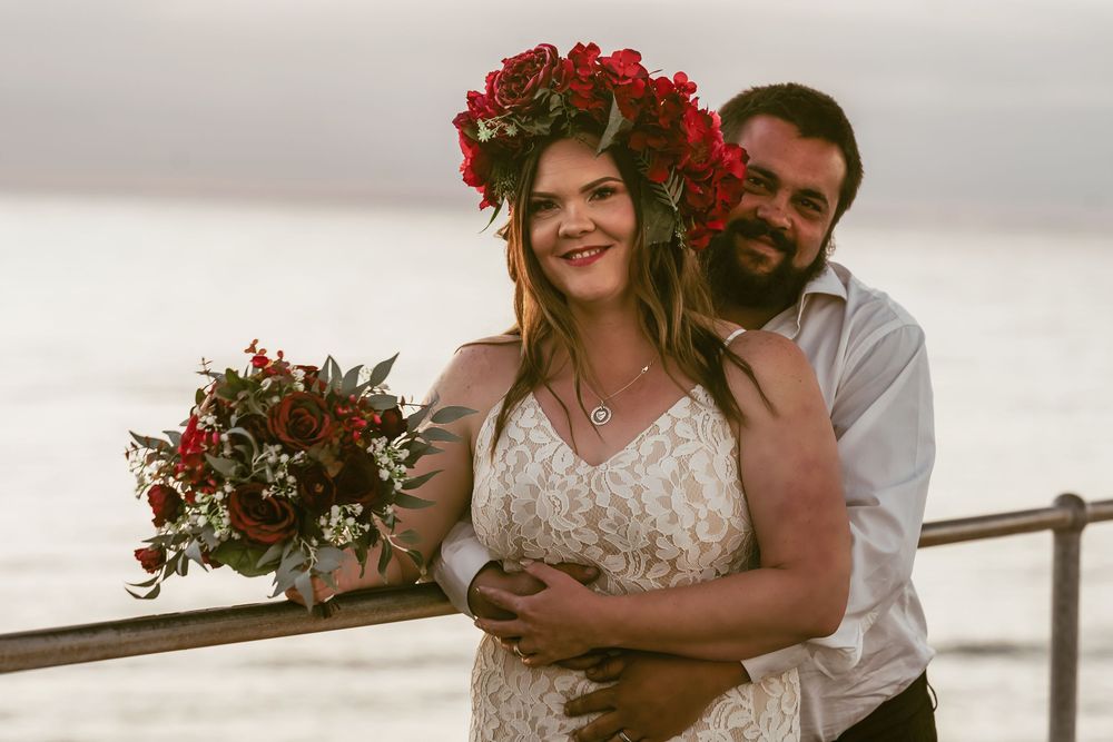 A couple in wedding attire embraces on a pier at sunset, with the bride wearing a floral crown and holding a rustic bouquet.