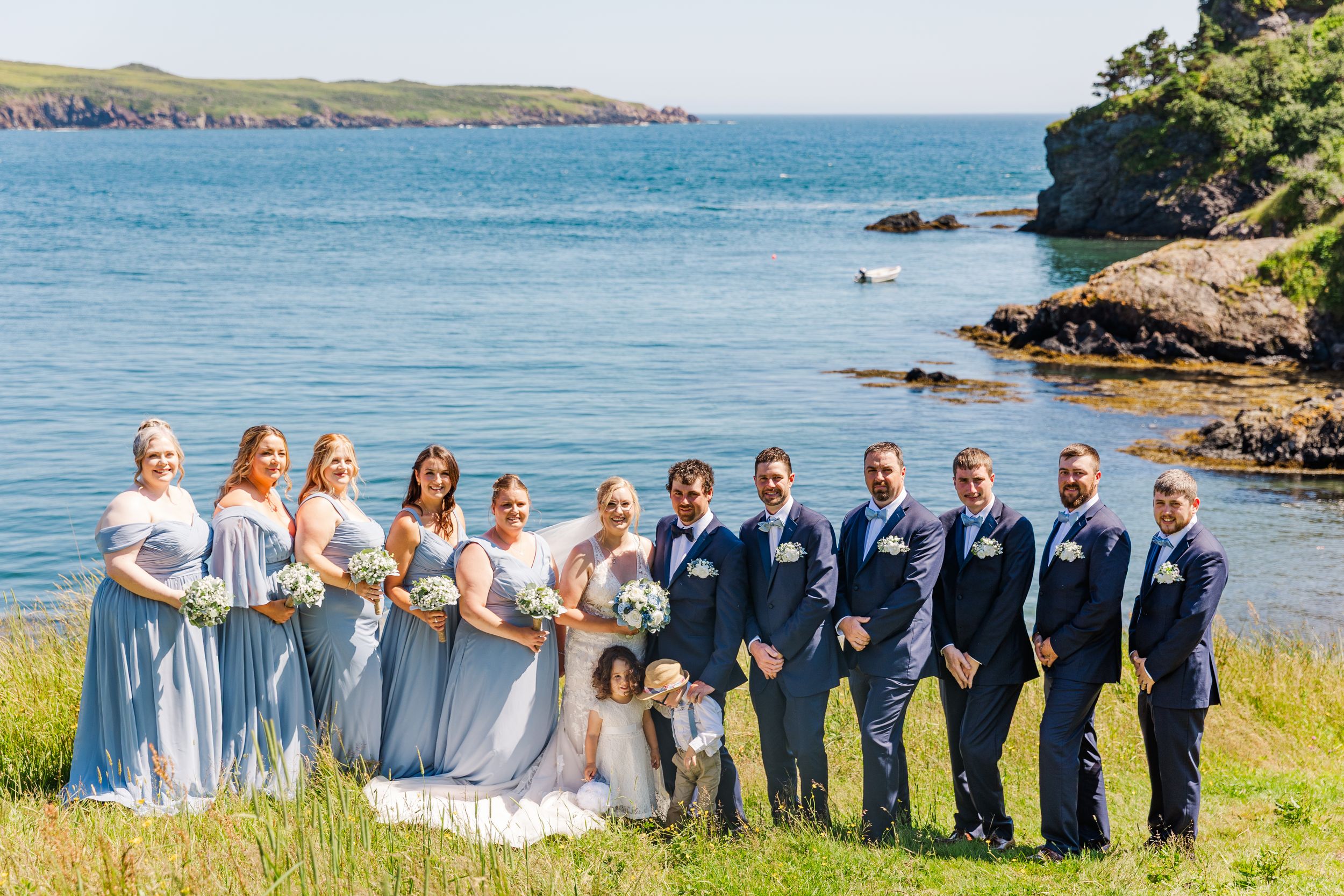 Wedding party poses on scenic coastal cliff with blue ocean and green hills in the background.