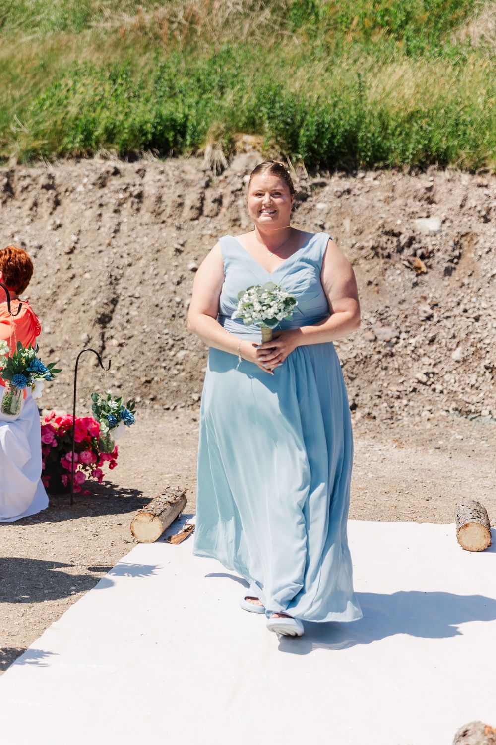 Person in light blue bridesmaid dress holding bouquet walks down white aisle runner at outdoor wedding ceremony.