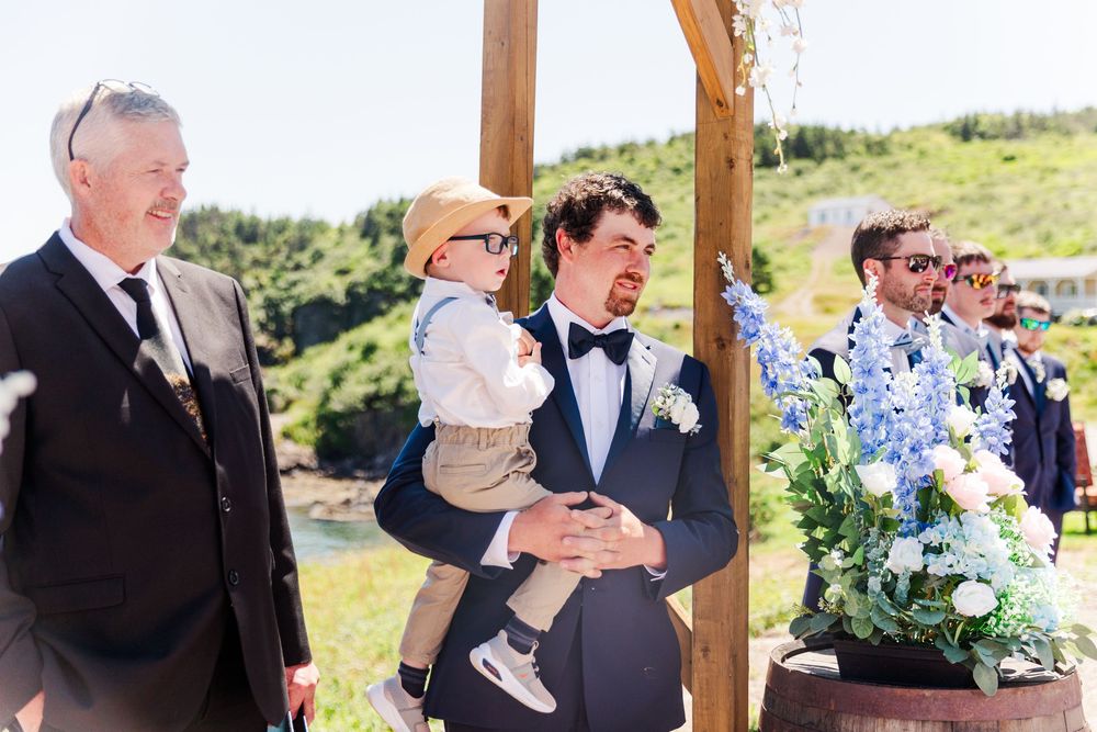 A wedding celebration takes place outdoors under a wooden pergola on a sunny hillside with guests gathered for the ceremony.
