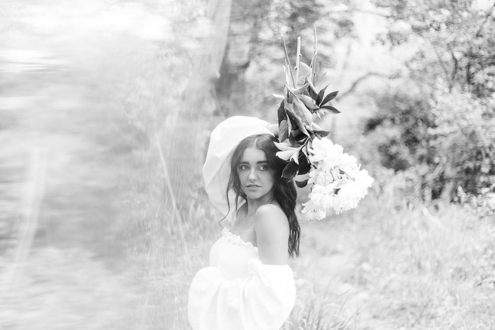 Ethereal black and white portrait of a woman in a white dress holding flowers in a dreamy outdoor setting.