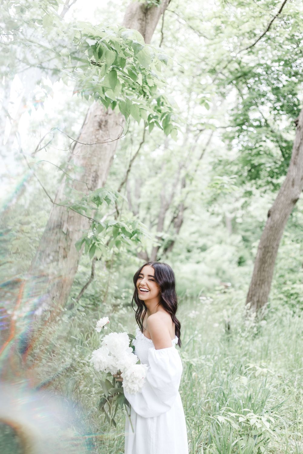 A person wearing a white top laughs joyfully while posing in a sunlit woodland setting with lush green foliage.