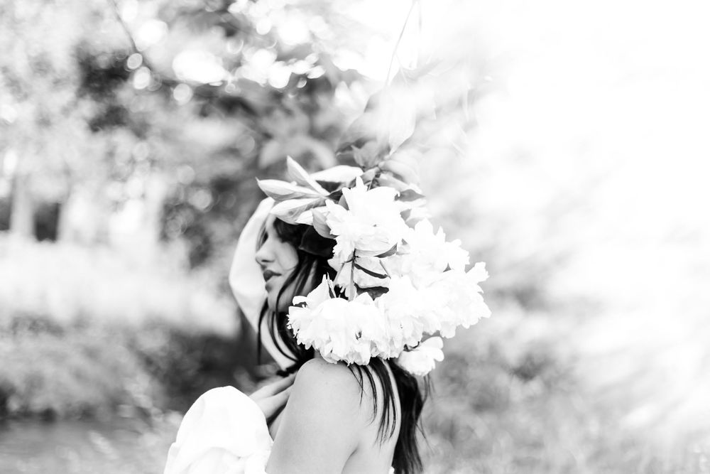 A black and white side profile portrait featuring a delicate floral crown headpiece in a dreamy outdoor setting.