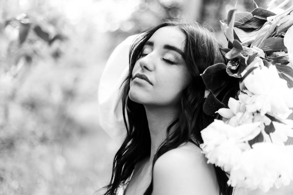 Black and white portrait of a young woman with closed eyes surrounded by delicate white flowers in nature.