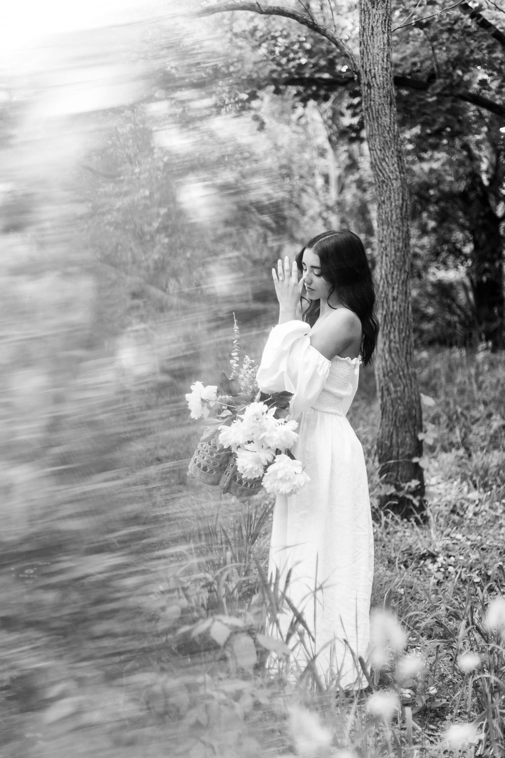 A dreamy black and white portrait of someone in a white off-shoulder dress holding flowers in a forest setting.