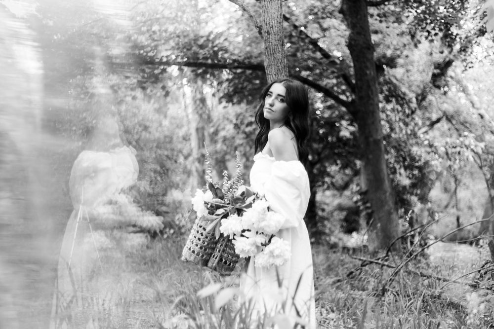Artistic black and white photograph showing a dreamy figure in white dress holding a basket in a sunlit forest setting.
