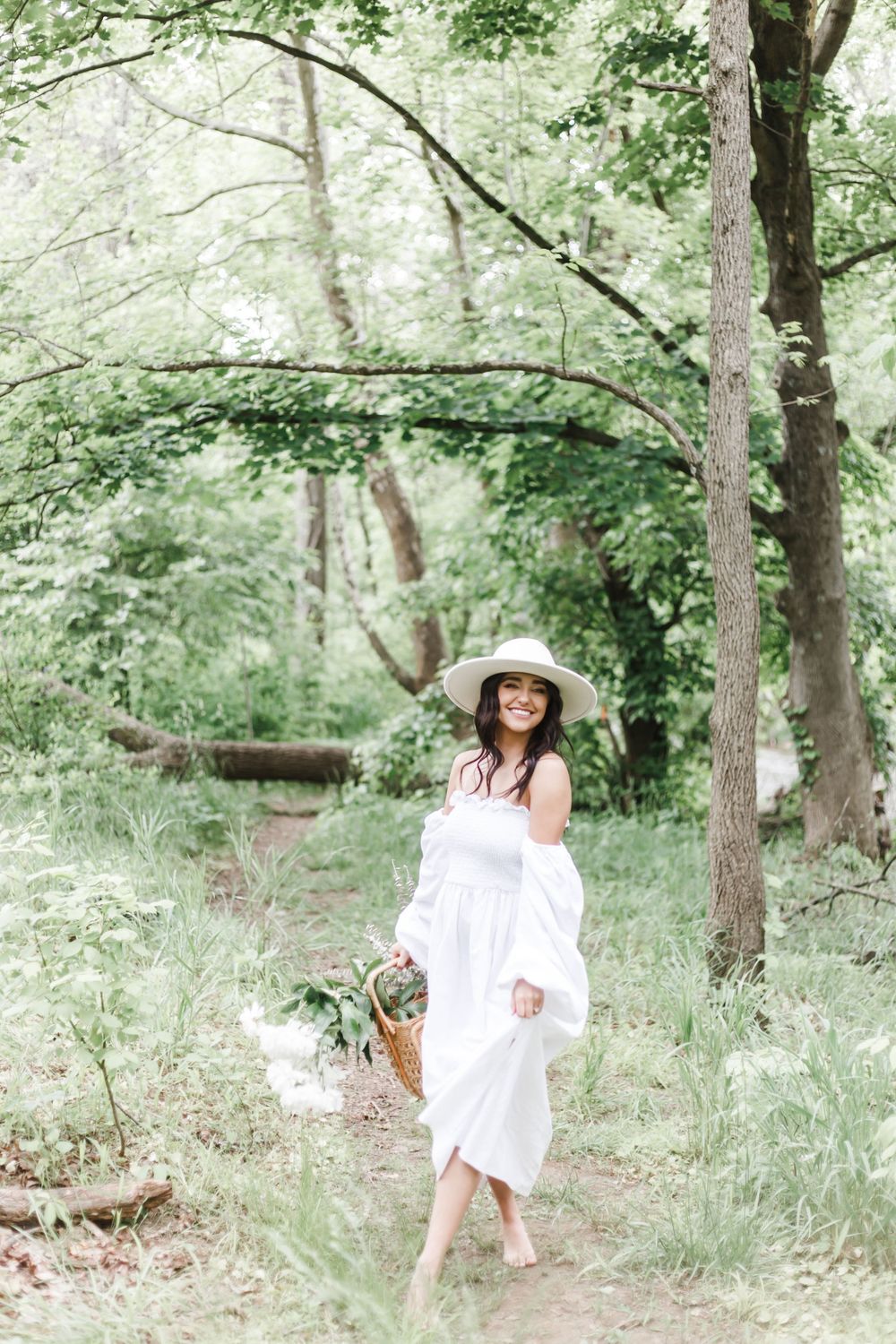 A dreamy summer photo session in a sun-dappled forest with someone in a white dress and straw hat.
