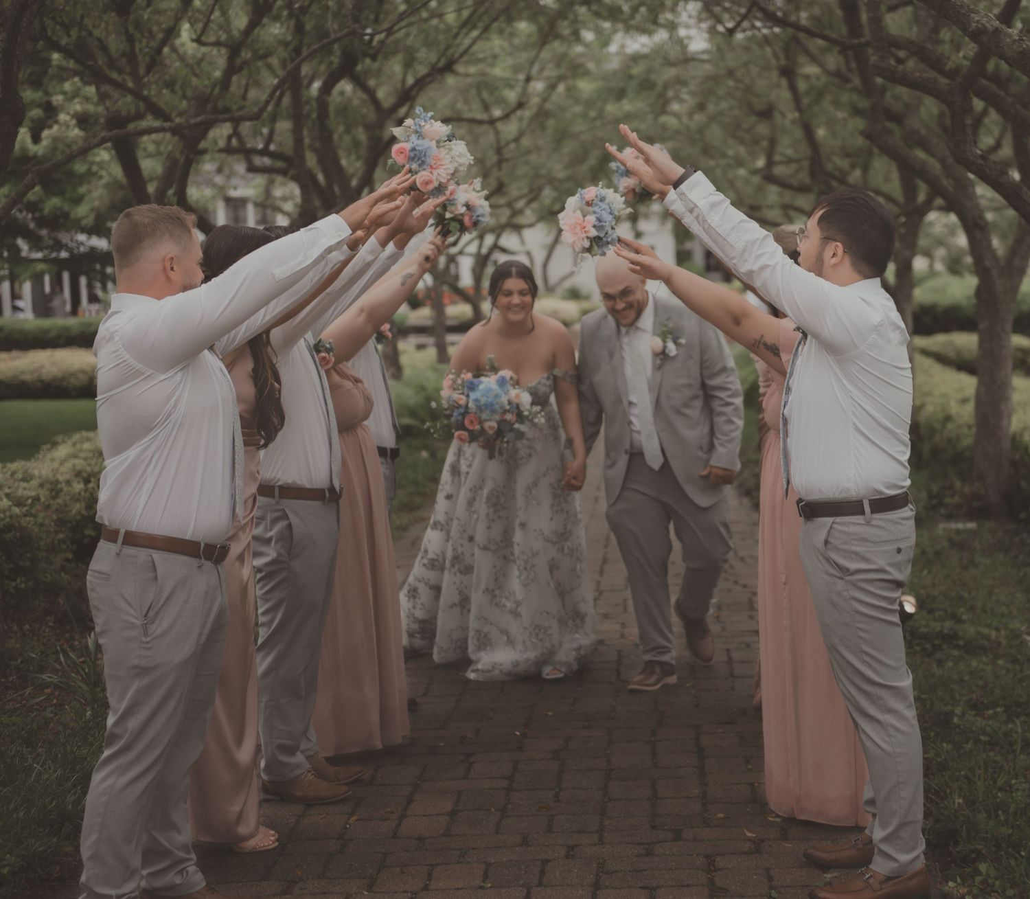 A wedding party performs a high five celebration tunnel on a brick pathway in a garden setting.