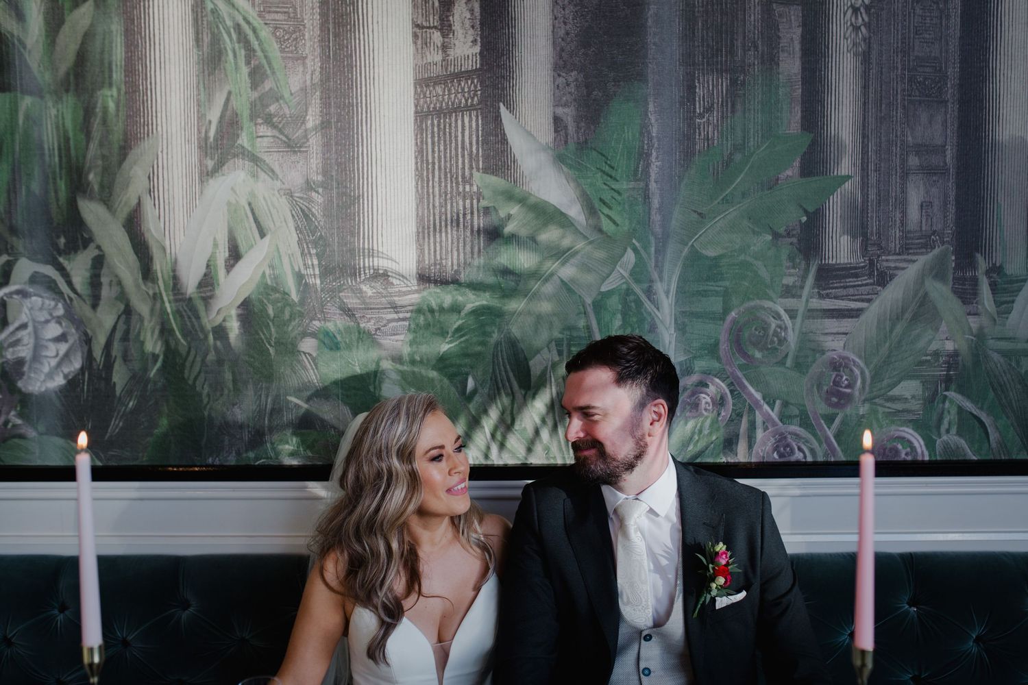 A couple shares a candid moment while seated together with lit candles and tropical plants behind them.
