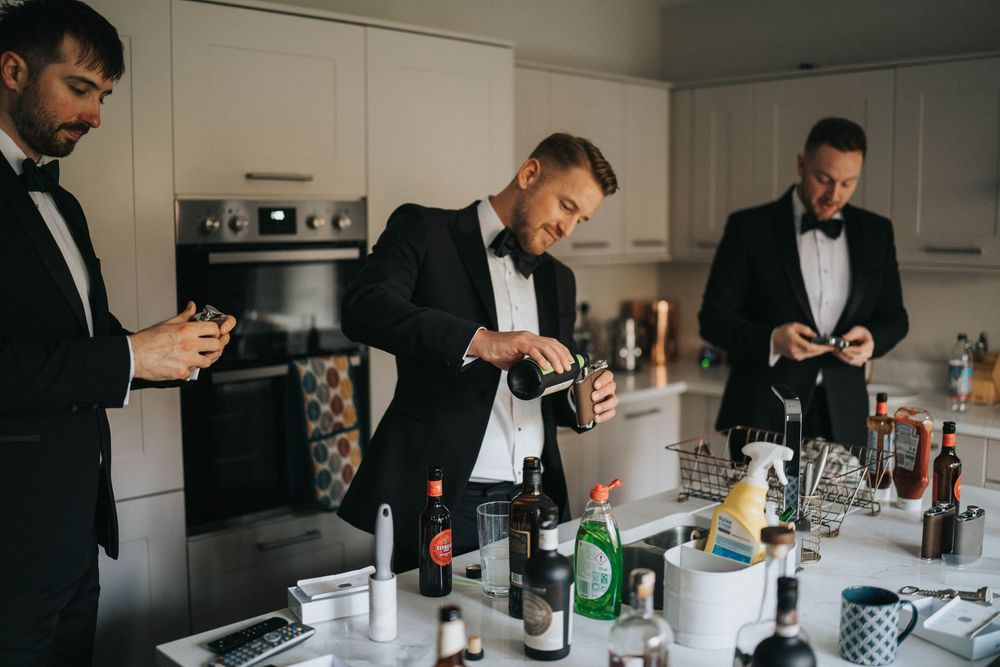 Groomsmen in black suits getting ready in kitchen with drinks and bottles on counter in moody lighting.