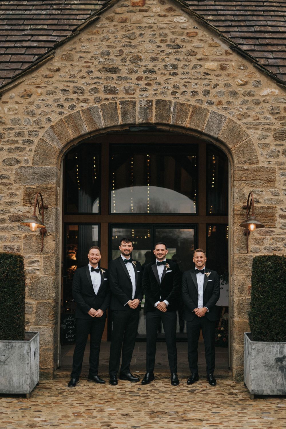 Groomsmen in black tuxedos stand in front of rustic stone archway entrance at elegant wedding venue.