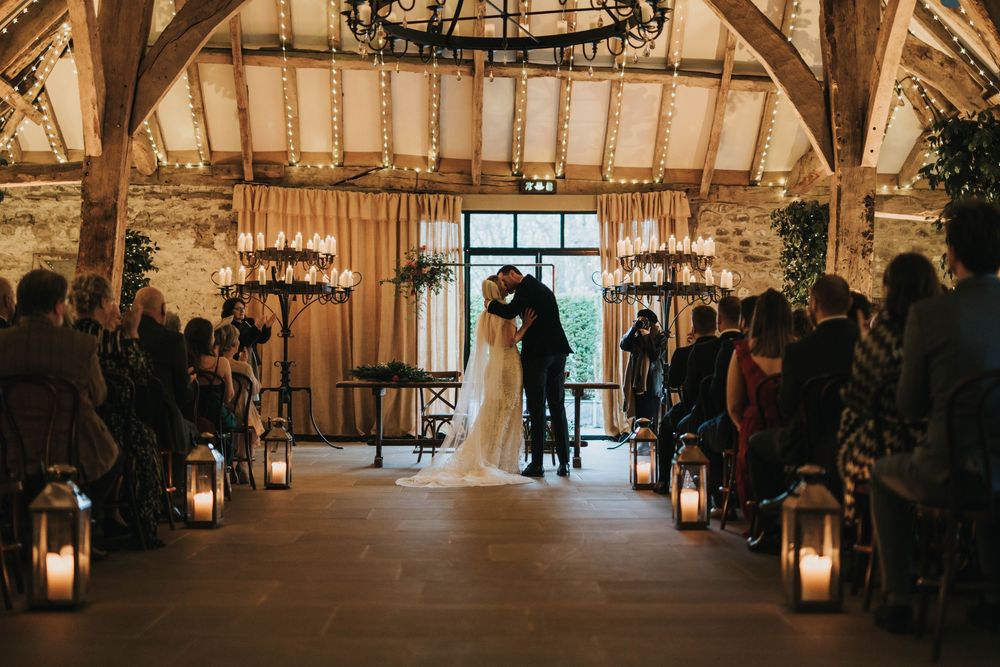 A rustic barn wedding ceremony with lantern-lined aisle and sunlight streaming through large windows behind the altar.