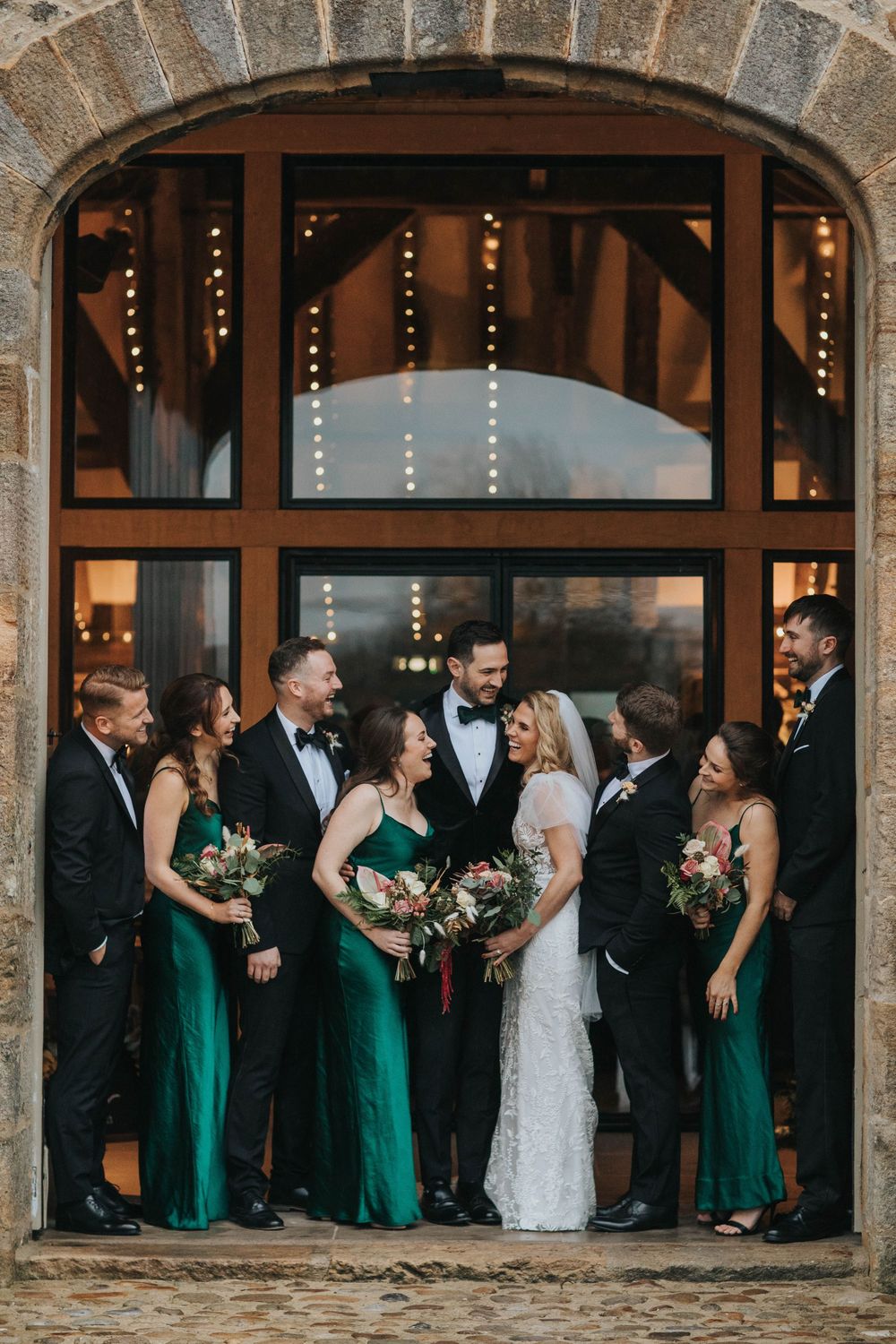 Wedding party in emerald green bridesmaid dresses and black tuxedos pose together under stone archway with twinkle lights.