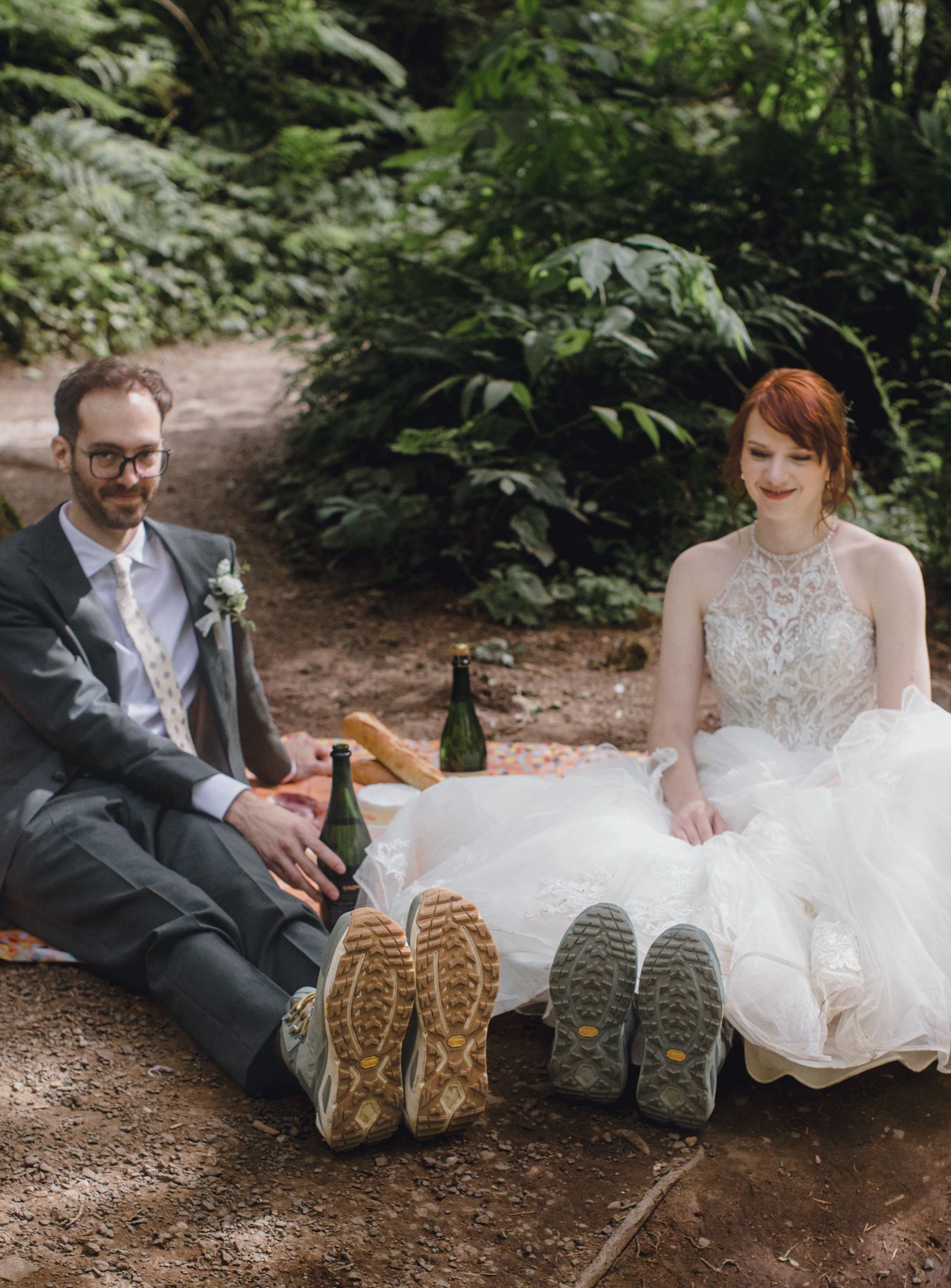 A hiking-boot-wearing couple sits comfortably on a forest trail with their wedding attire and bottles of beer.