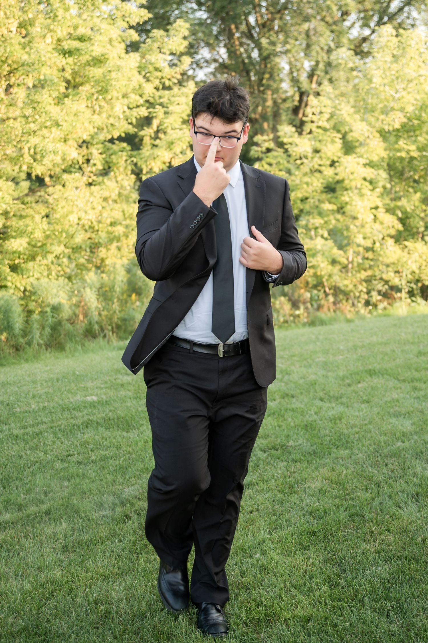 A person in a black suit and tie walks on green grass with yellow foliage in the background.