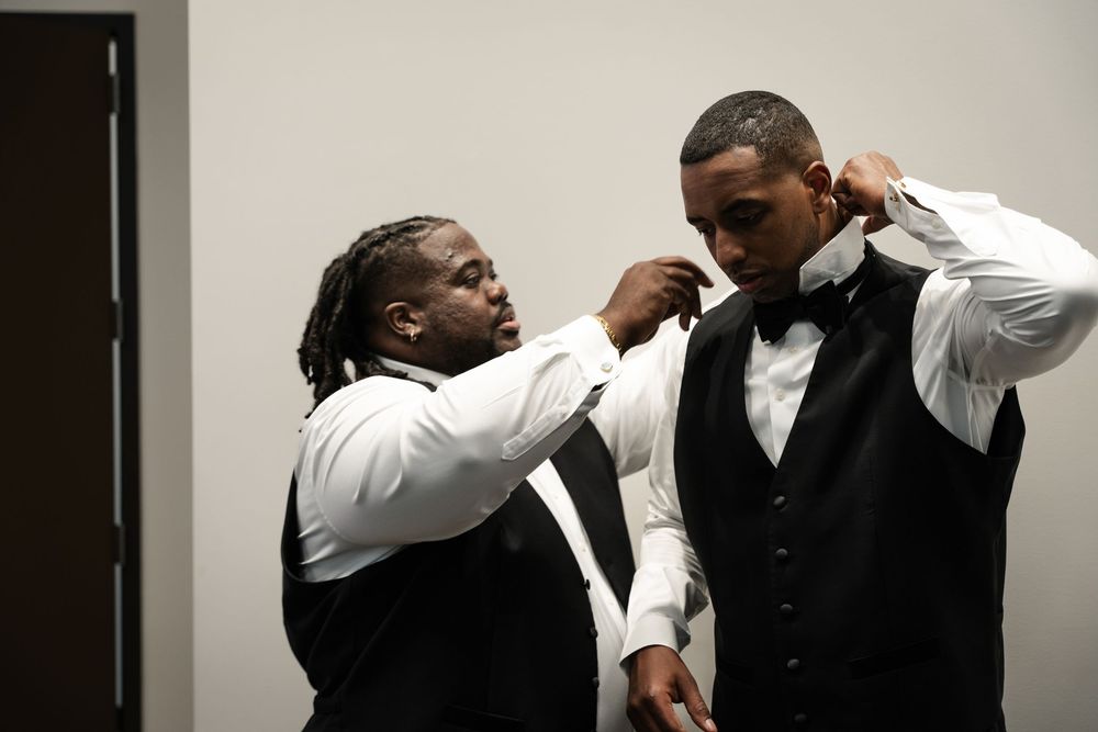 Two people in formal attire assist each other with adjusting bow ties before an event.
