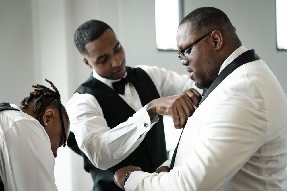 Groomsmen in white tuxedos share emotional embraces during pre-wedding preparations.