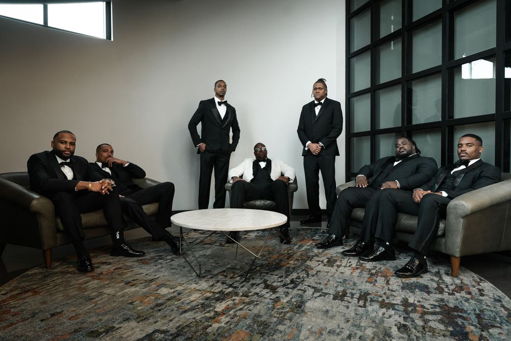 Group in formal black suits and ties sits on modern gray stone steps near glass windows during formal event.