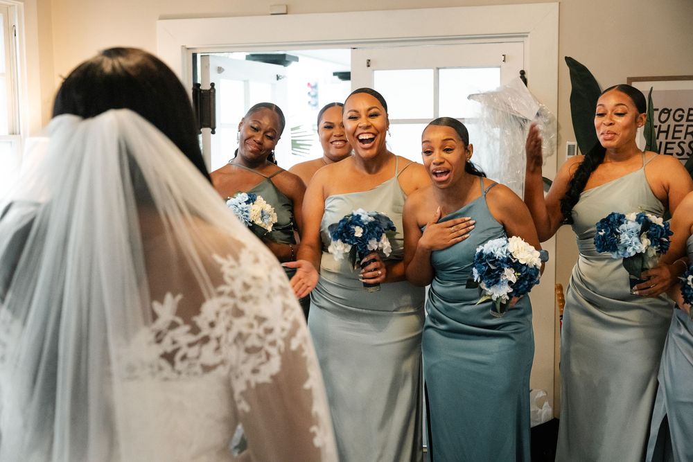 Bridesmaids in dusty blue dresses share joyful reactions during wedding preparations.