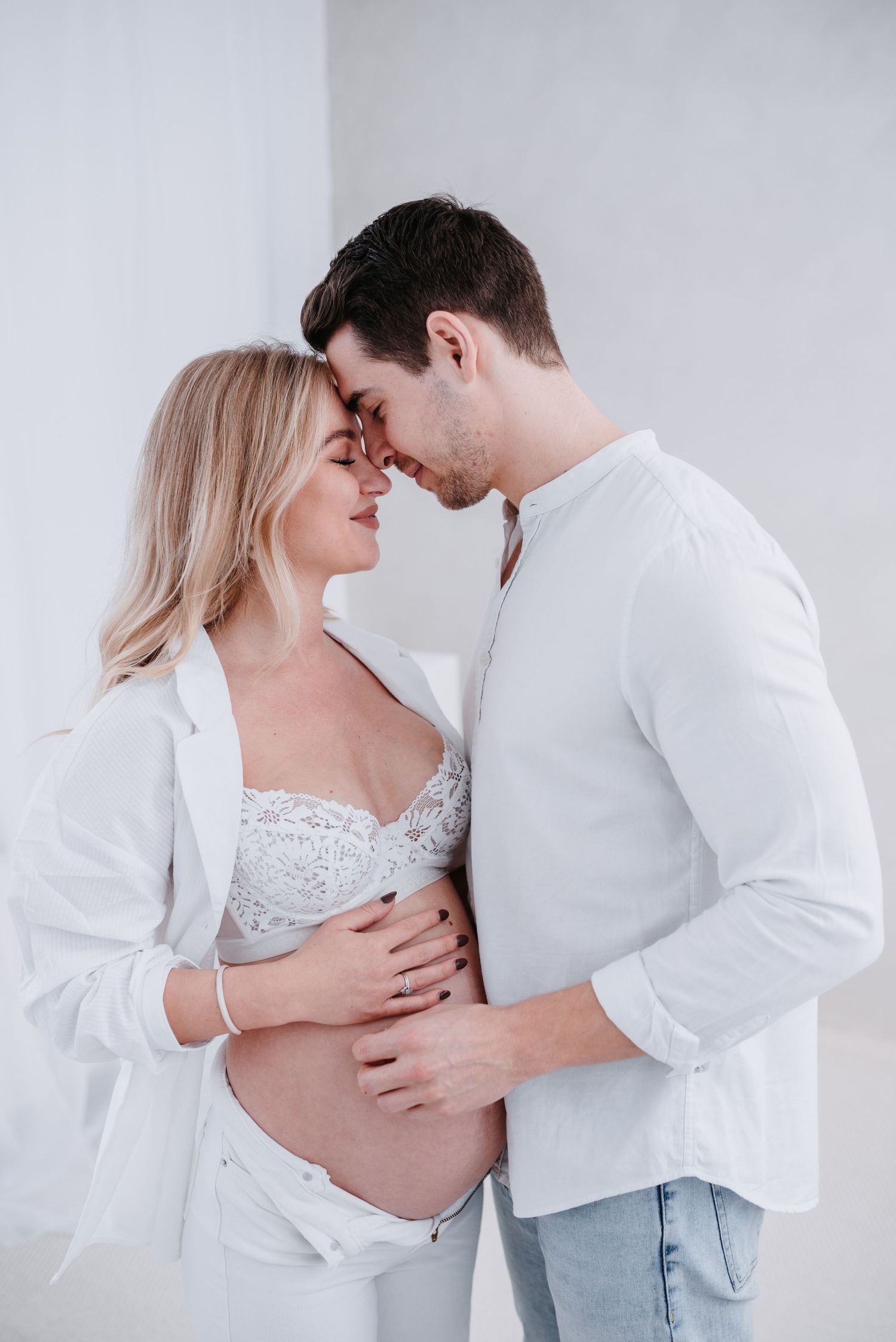 Elegant maternity photoshoot showing intimate moments between couple in white and gray attire against light background.
