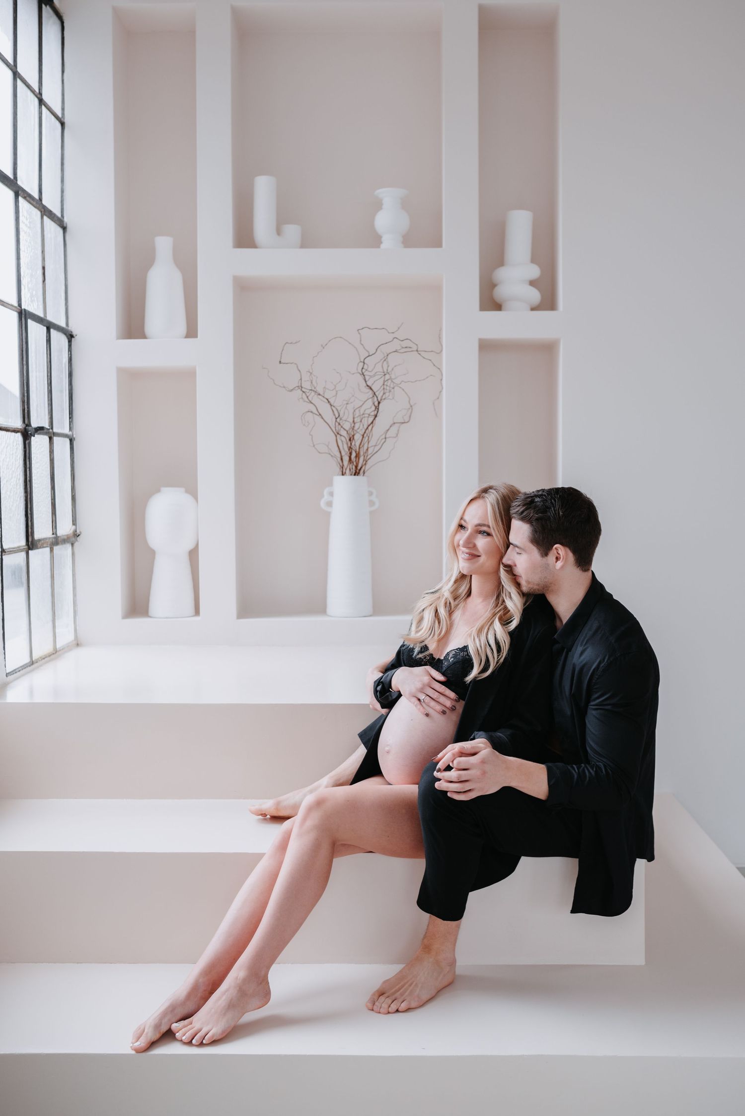 Romantic couple shares joyful moment sitting together on white minimalist steps near window with decorative display niches.