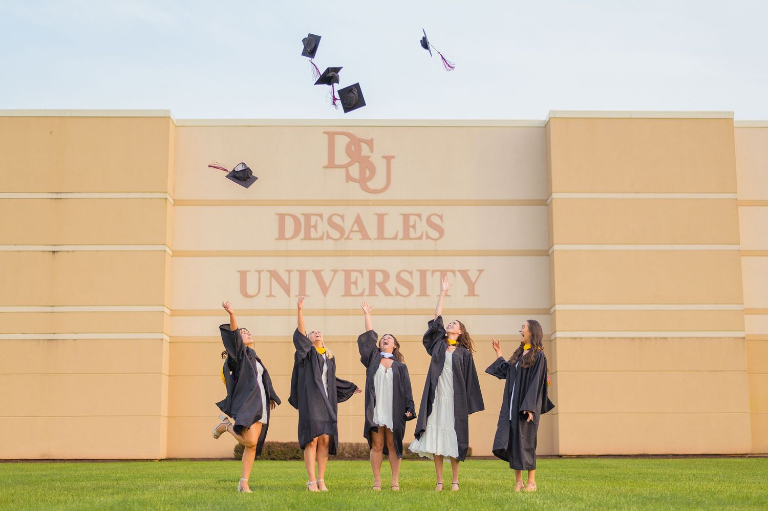 Graduates tossing their caps in the air in front of DeSales University building sign.