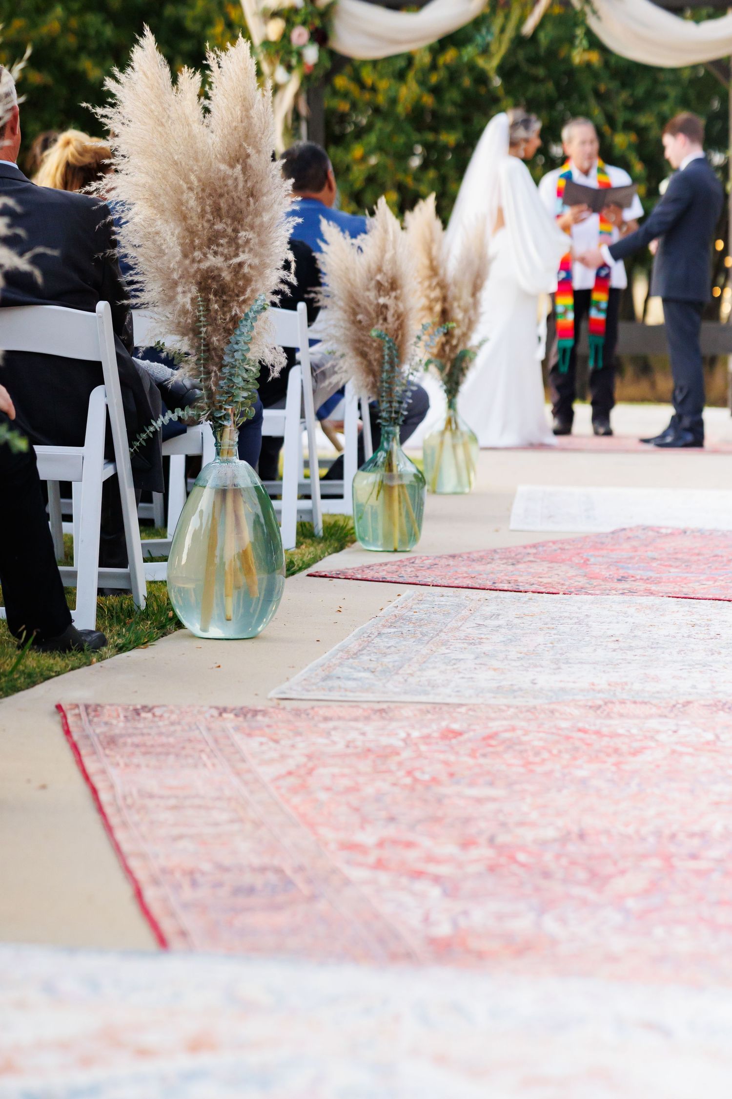 Decorative glass vases with pampas grass line an outdoor wedding ceremony aisle covered in pink patterned carpet.