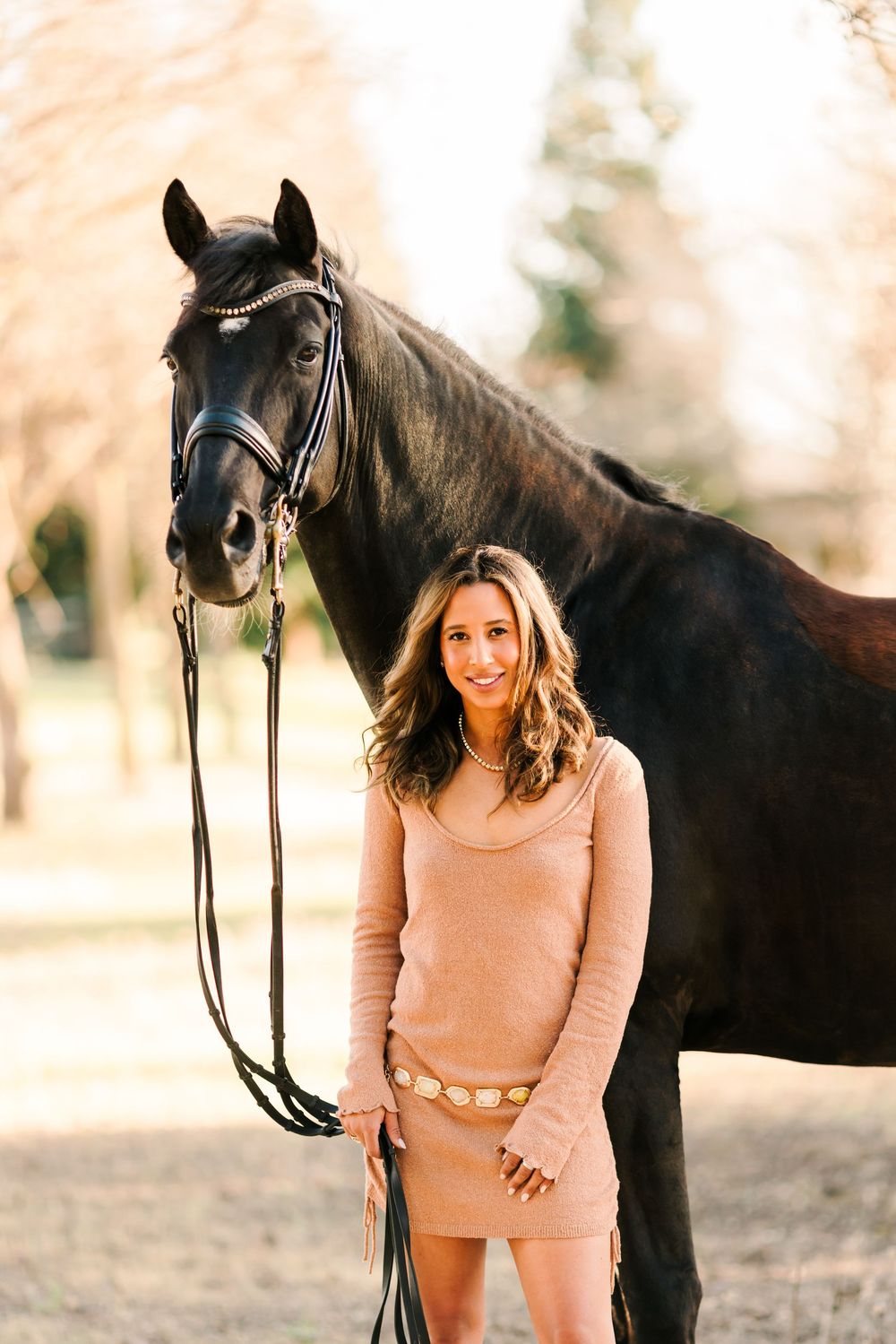 A dark horse wearing bridle and reins stands with someone in a pink sweater outdoors.