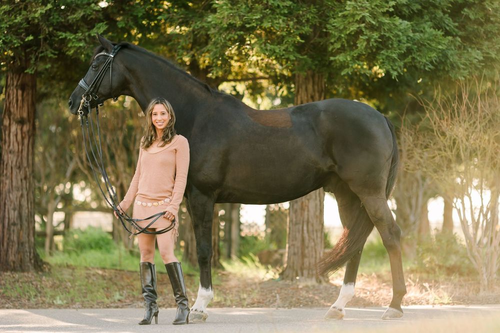 A dark horse stands on a path in dappled sunlight beside someone wearing a peach sweater and riding boots.