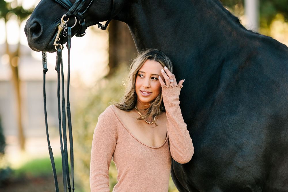 A dark horse wearing riding tack stands next to someone in casual attire outdoors.