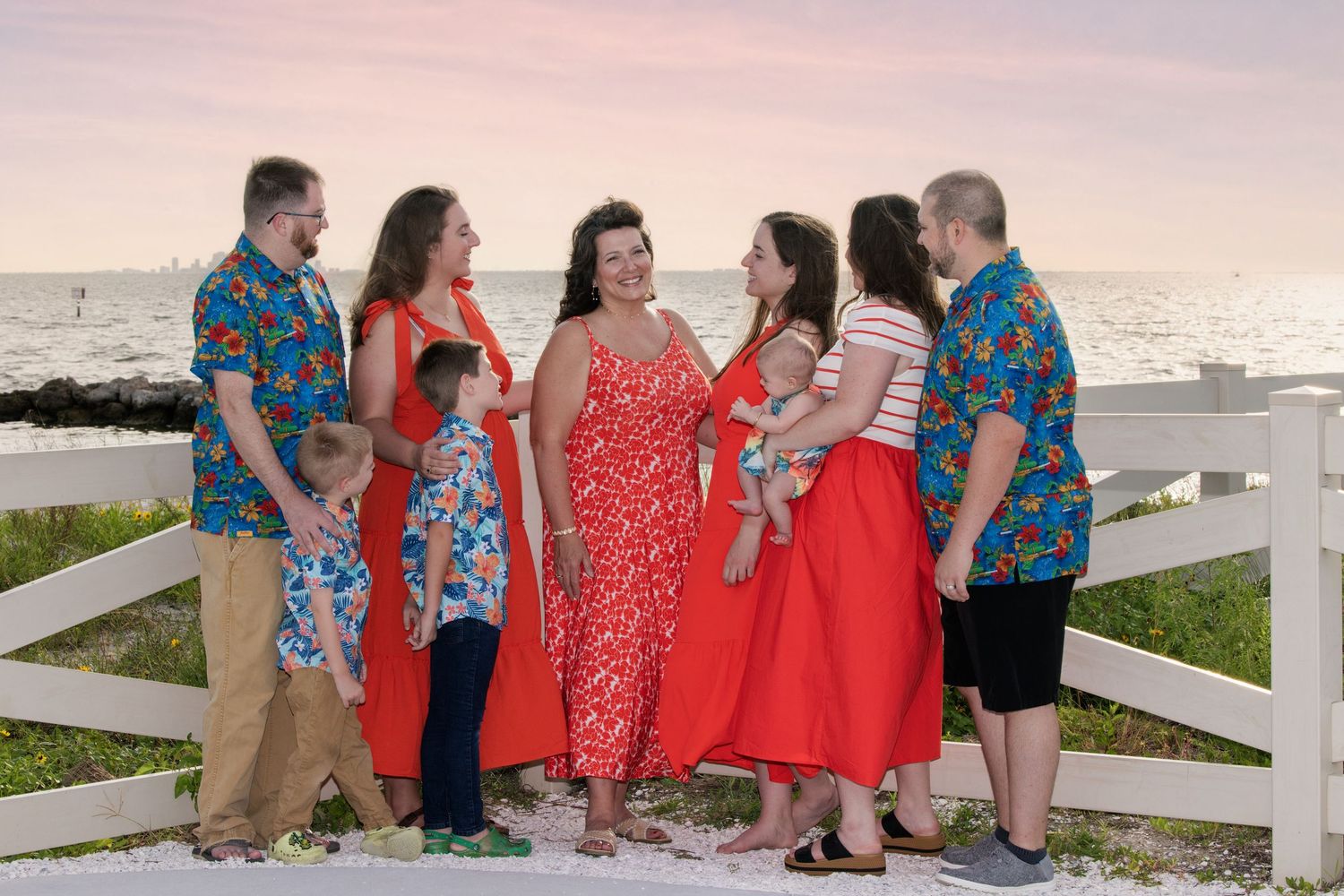 A family group in coordinating red and blue outfits poses together at sunset by a white fence near the beach.