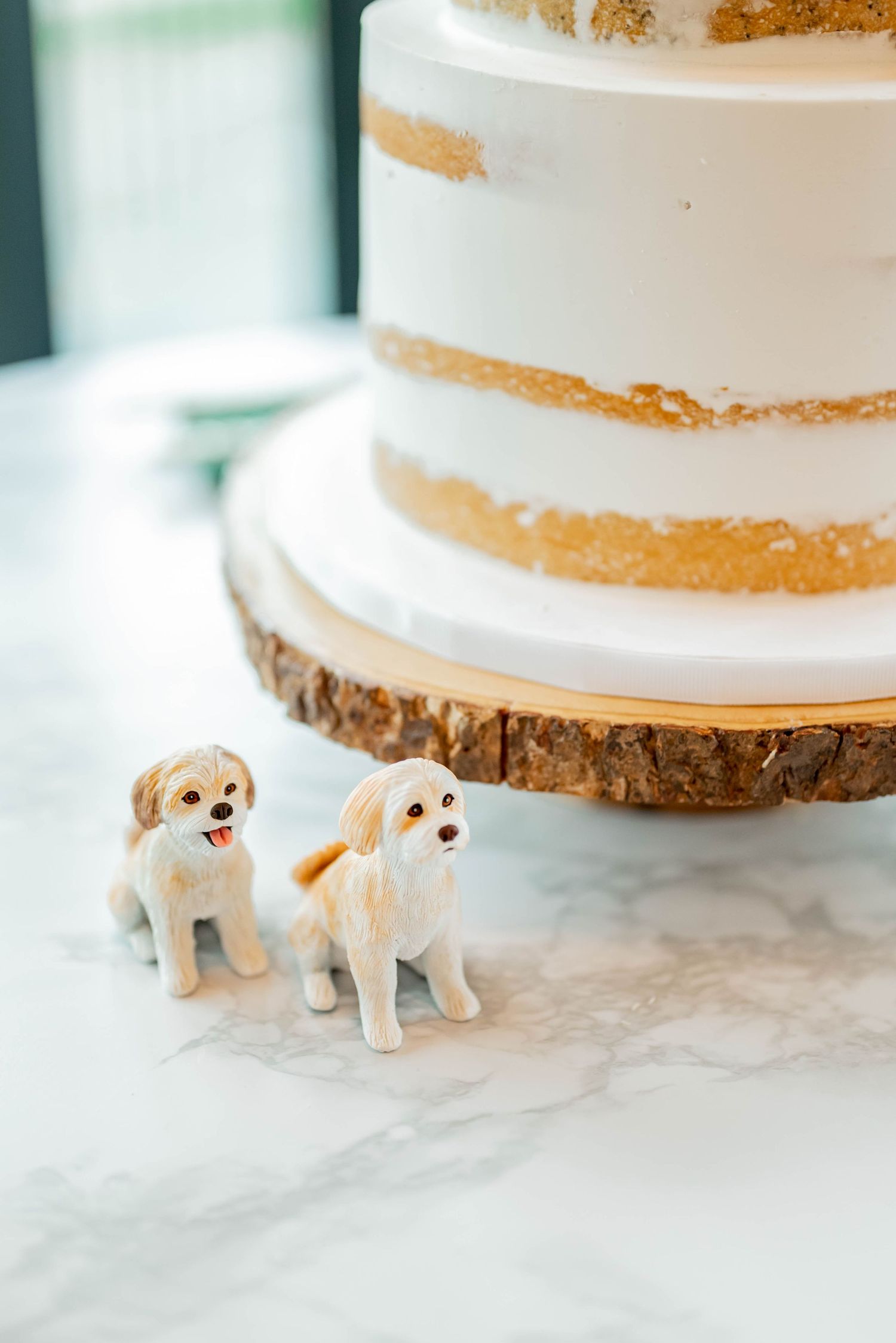 Two small white clay or fondant dog cake toppers sitting on a table next to a wedding cake with rustic wooden base.