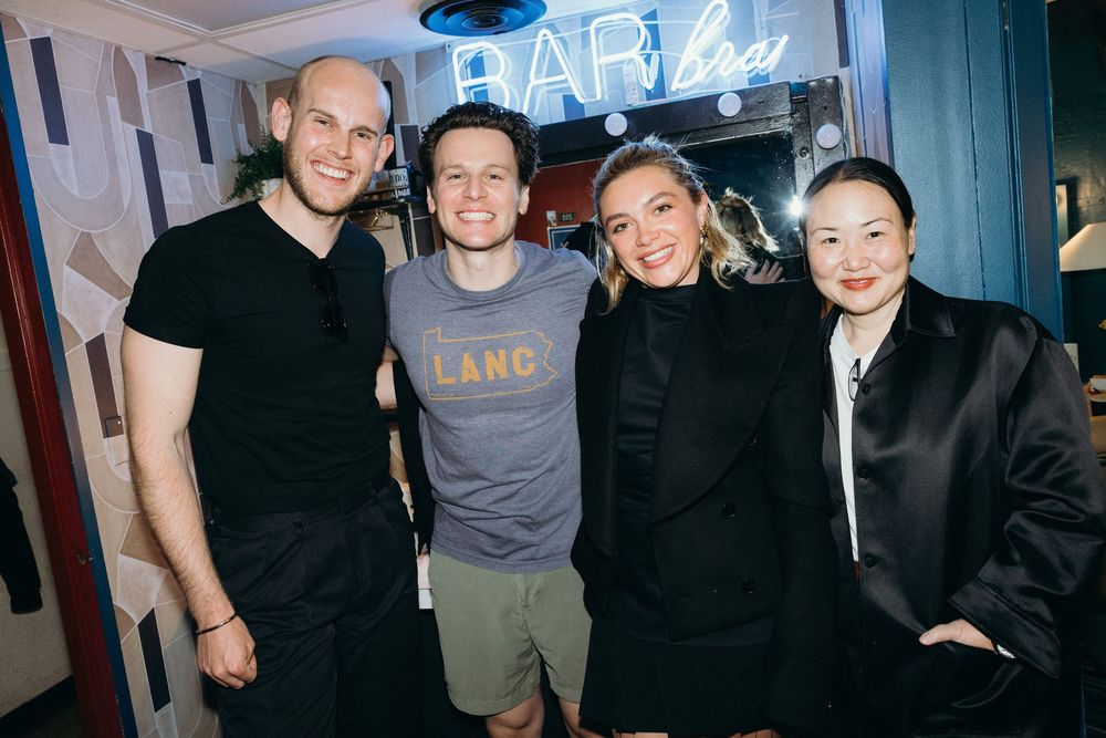 Four people stand together smiling in front of a blue neon sign at what appears to be a bar or venue.