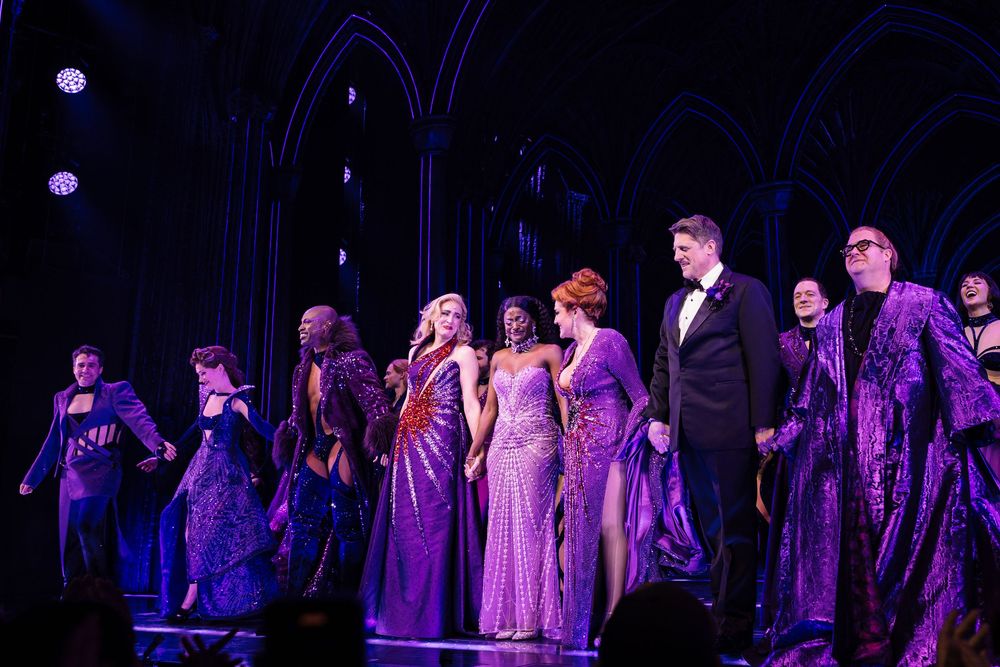 A group of performers takes their final bow on a dark stage illuminated by purple lighting during a theatrical show.