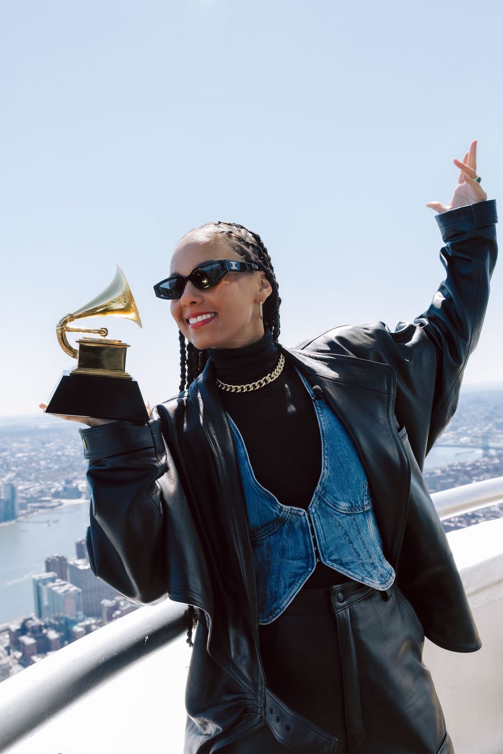 Person in sunglasses and denim vest holds up a Grammy Award trophy while posing on a rooftop overlooking a cityscape.