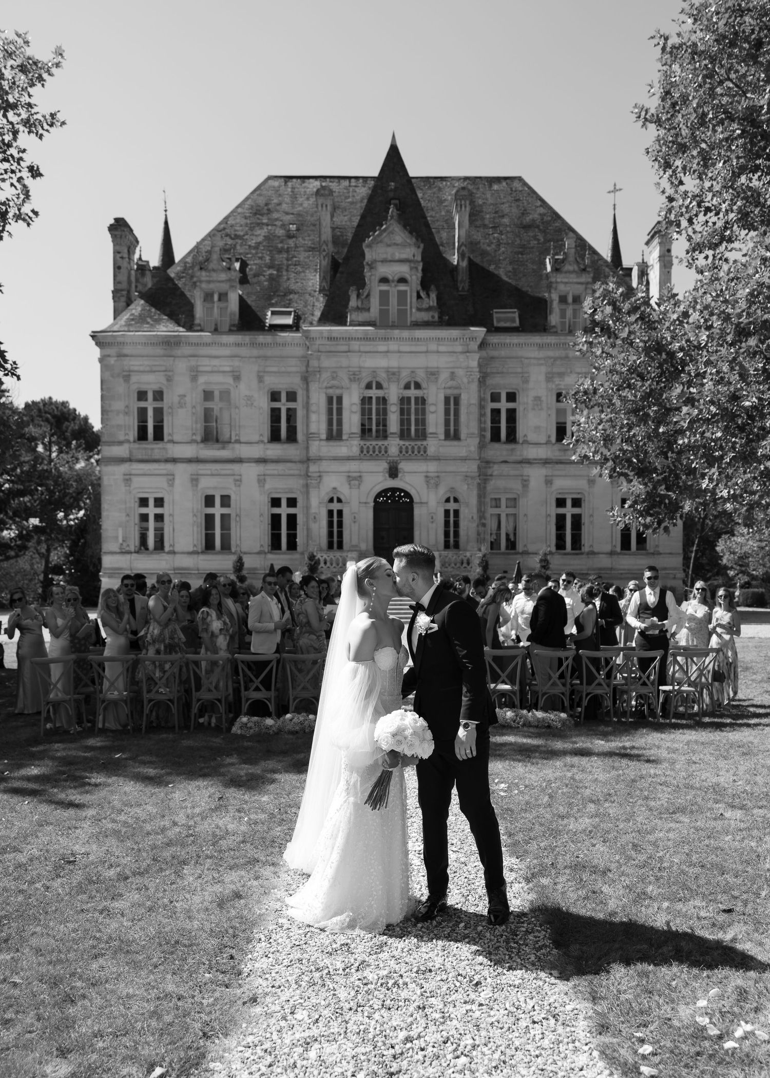 A black and white wedding photo in front of a grand chateau with a crowd of guests standing in background.