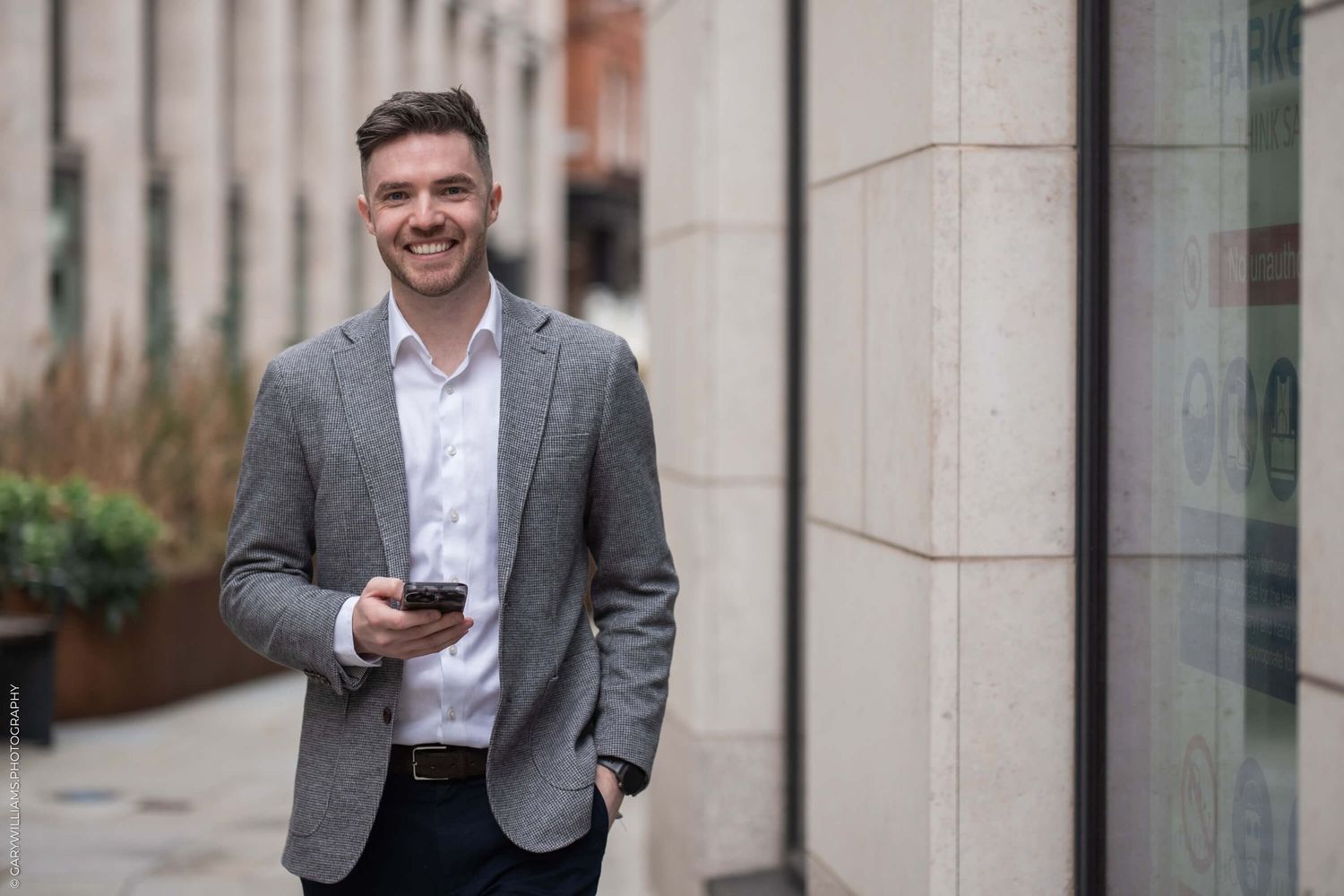 Professional in grey blazer and white shirt standing outside office building holding smartphone with friendly expression.