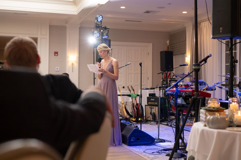 A performer in a lavender dress sings on stage at an elegant indoor venue with professional lighting and musical equipment.