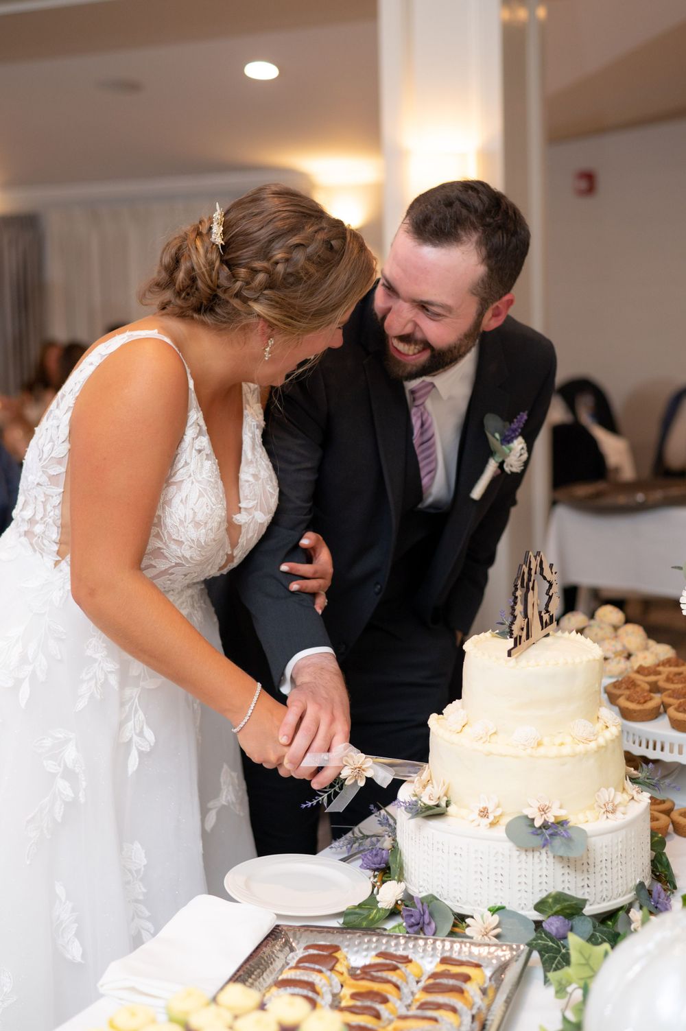 Happy newlyweds cut their elegant white wedding cake together at the reception while sharing a laugh.