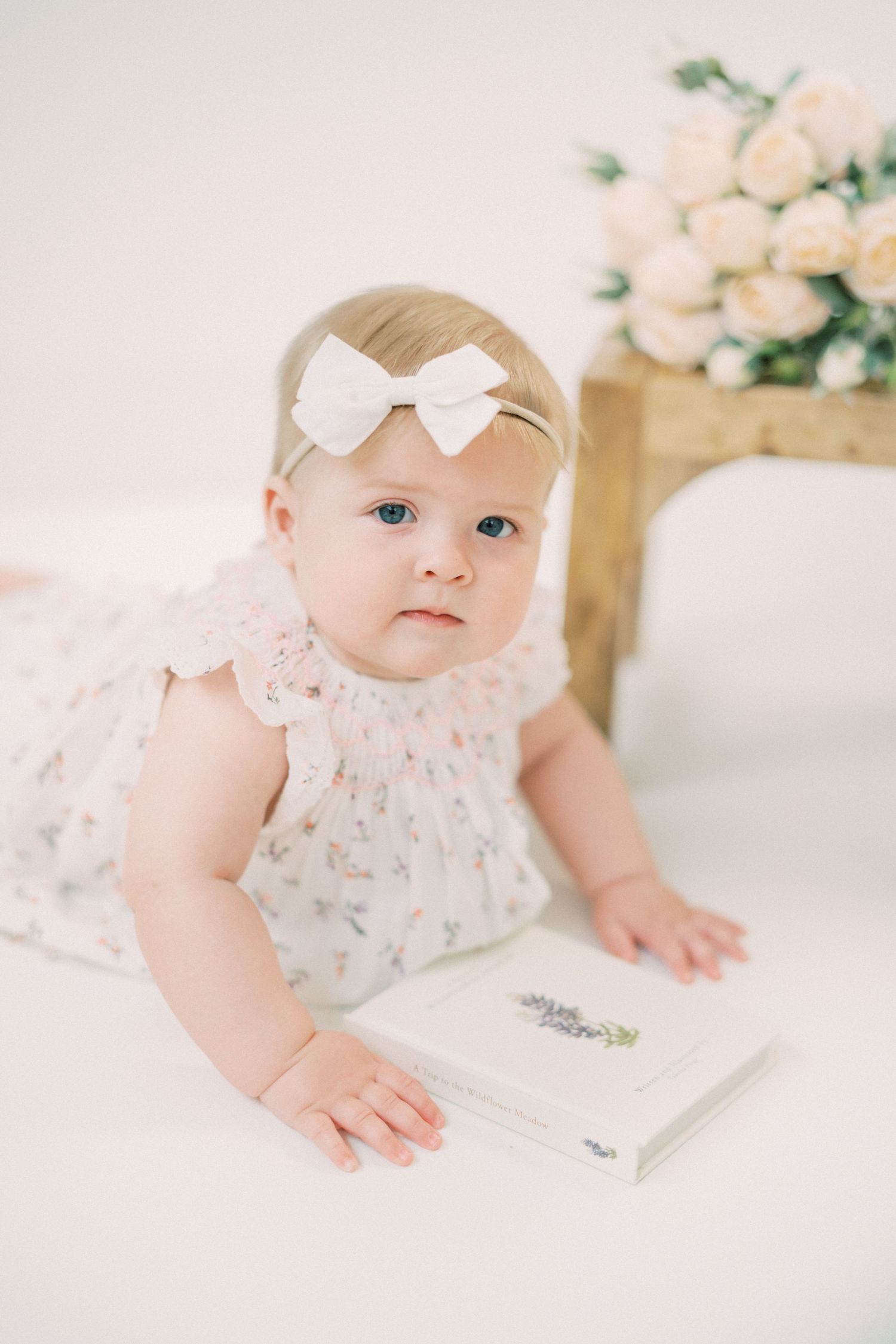 Sweet baby in floral dress with white bow headband lies on soft bedding next to a book and white roses in background.